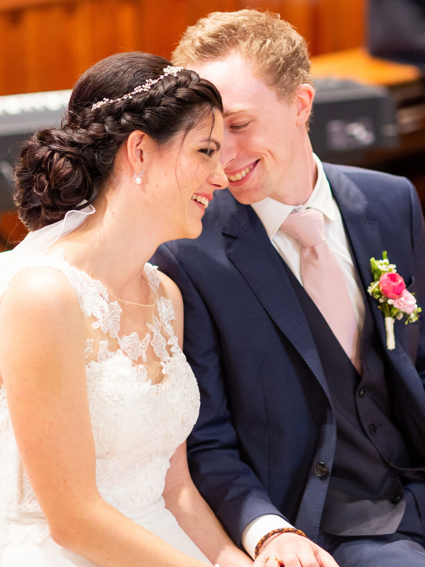 Ein intimer Moment zwischen Braut und Bräutigam während ihrer Trauung. Die Braut in einem weißen, spitzenbesetzten Kleid lehnt sich lächelnd zu ihrem Bräutigam, der einen dunklen Anzug und eine rosa Krawatte trägt. Beide strahlen vor Glück.