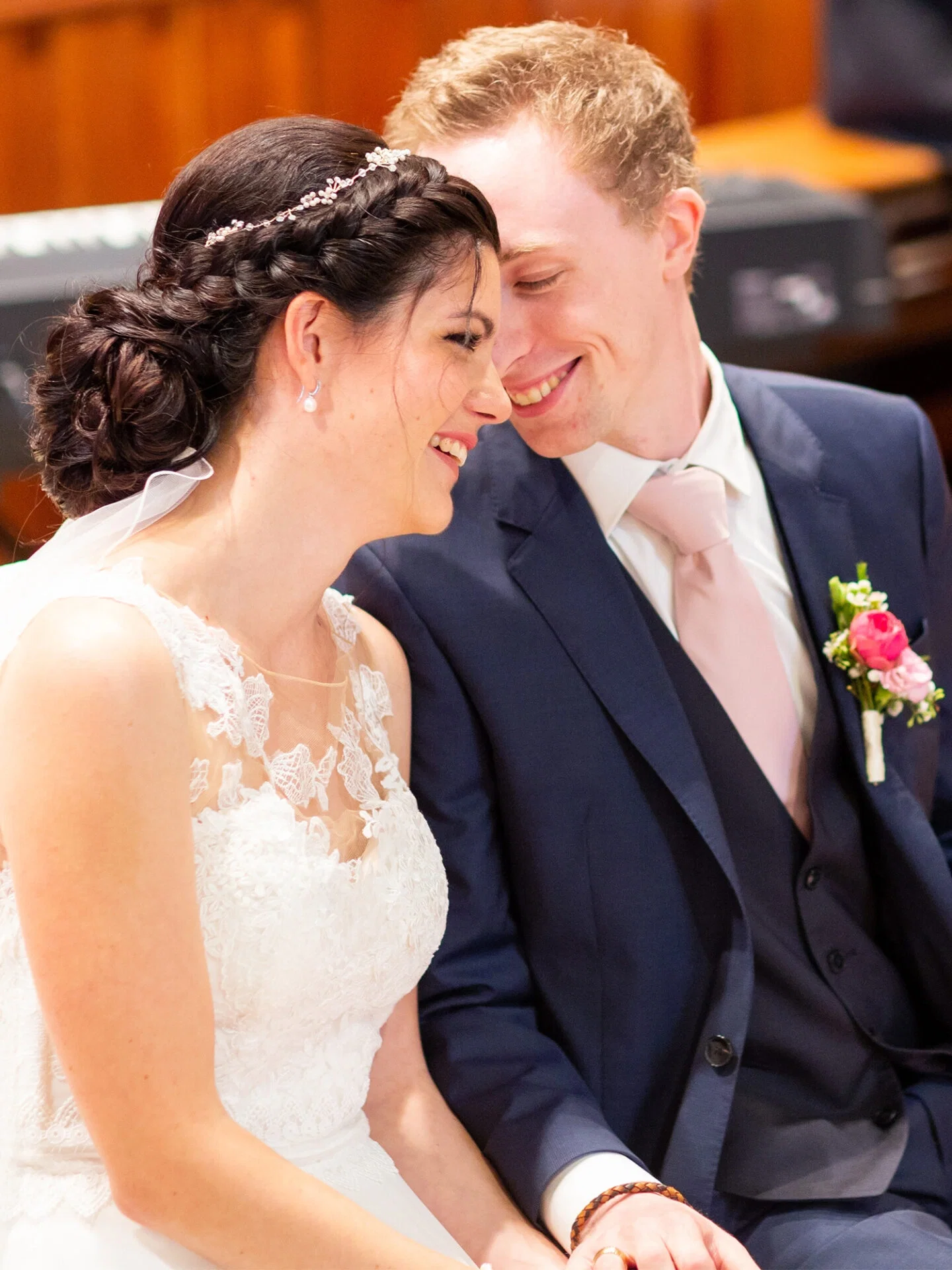 Ein intimer Moment zwischen Braut und Bräutigam während ihrer Trauung. Die Braut in einem weißen, spitzenbesetzten Kleid lehnt sich lächelnd zu ihrem Bräutigam, der einen dunklen Anzug und eine rosa Krawatte trägt. Beide strahlen vor Glück.