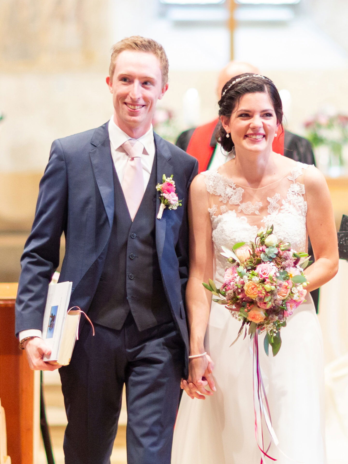 Das glückliche Paar geht nach der Hochzeitszeremonie Arm in Arm durch die Kirche. Die Braut trägt ein Spitzenkleid und hält einen frischen Blumenstrauß, während der Bräutigam mit einem Buch in der Hand lächelnd an ihrer Seite geht.