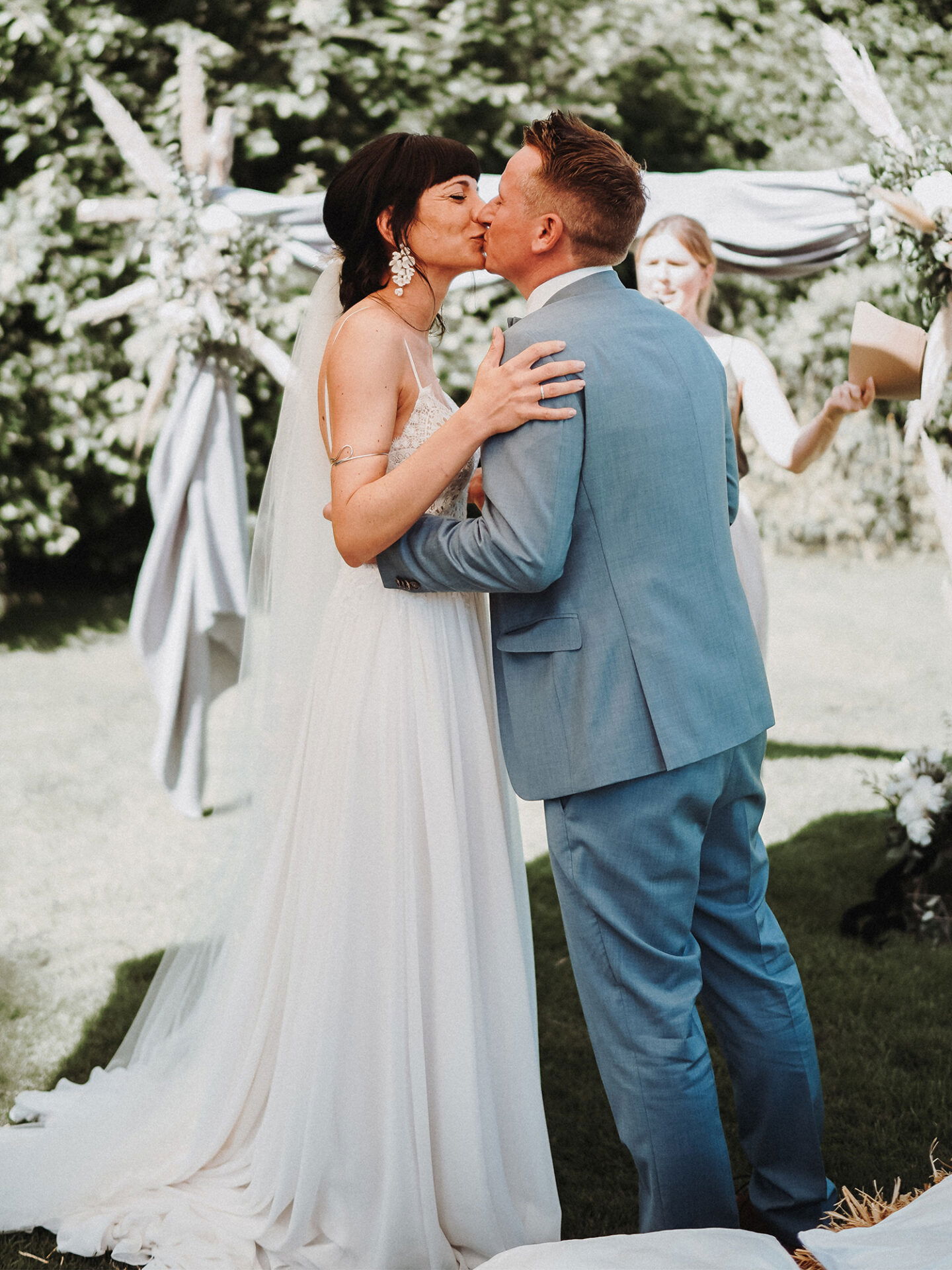 Ein verliebtes Brautpaar teilt einen Kuss bei ihrer Hochzeit im Freien. Die Braut in einem eleganten, fließenden Kleid und der Bräutigam in einem pastellblauen Anzug umarmen sich liebevoll. Im Hintergrund steht eine Rednerin bereit.