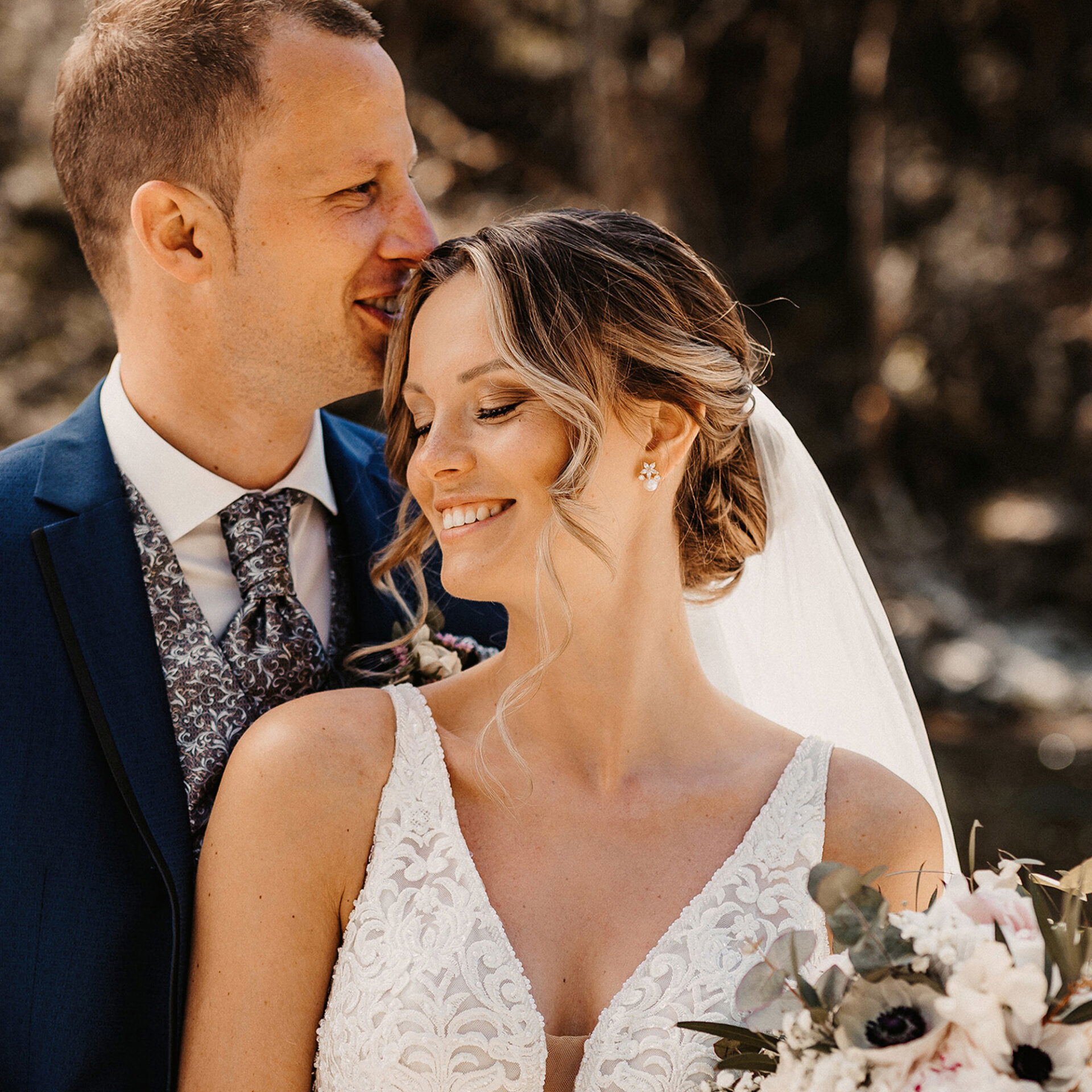 Das Brautpaar genießt einen intimen Moment; die Braut lächelt sanft, während der Bräutigam sie liebevoll anschaut, umgeben von natürlichem Tageslicht.