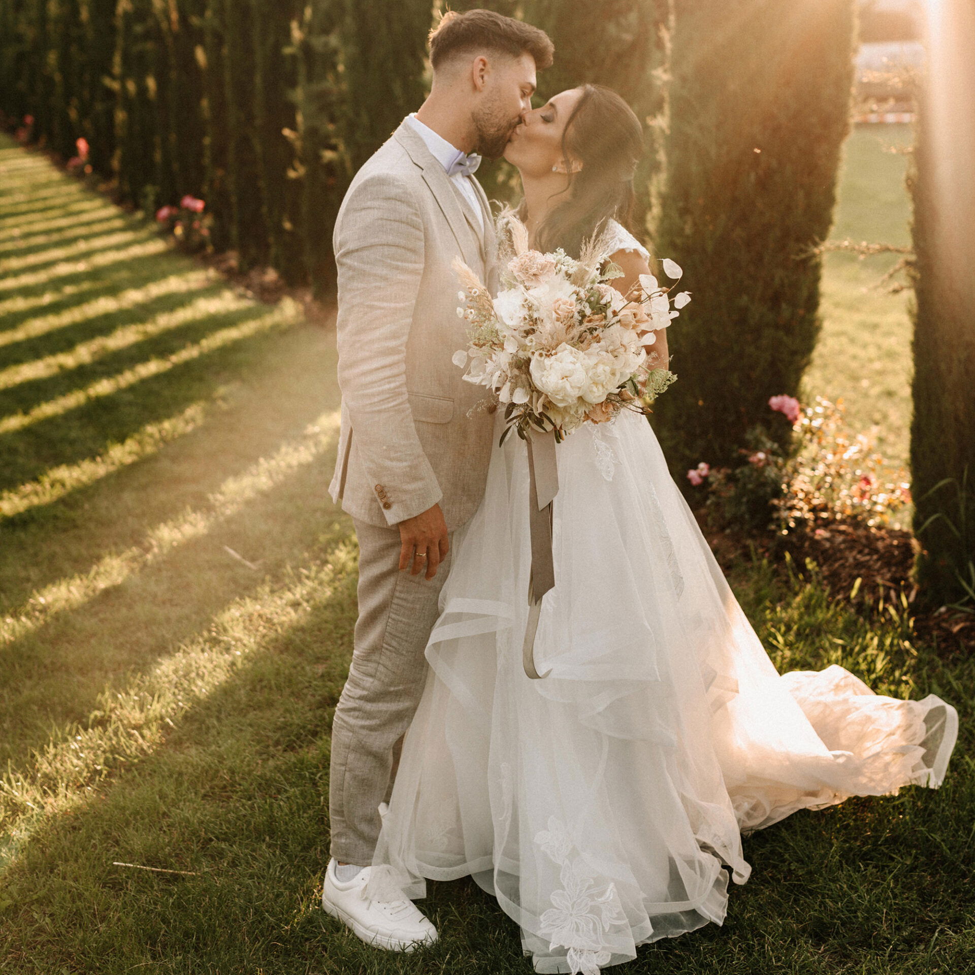 Das Brautpaar steht in einem von Bäumen gesäumten Garten und teilt einen romantischen Kuss im warmen Licht des Sonnenuntergangs. Die Braut trägt ein weißes Kleid mit Schleppe und der Bräutigam einen hellen Anzug, während die Sonnenstrahlen sanft durch die Bäume scheinen.