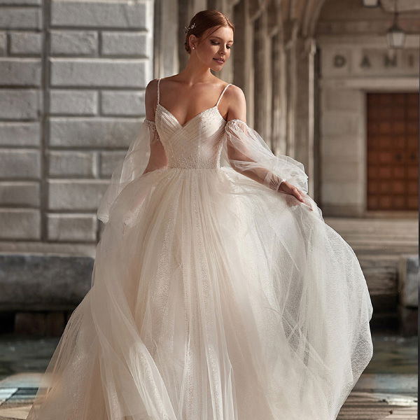 Eine Frau in einem weißen, schulterfreien Brautkleid posiert vor einem alten Gebäude mit Steinmauern. Der voluminöse Tüllrock ihres Kleides bewegt sich leicht, während sie elegant den Saum hält und anmutig dasteht.