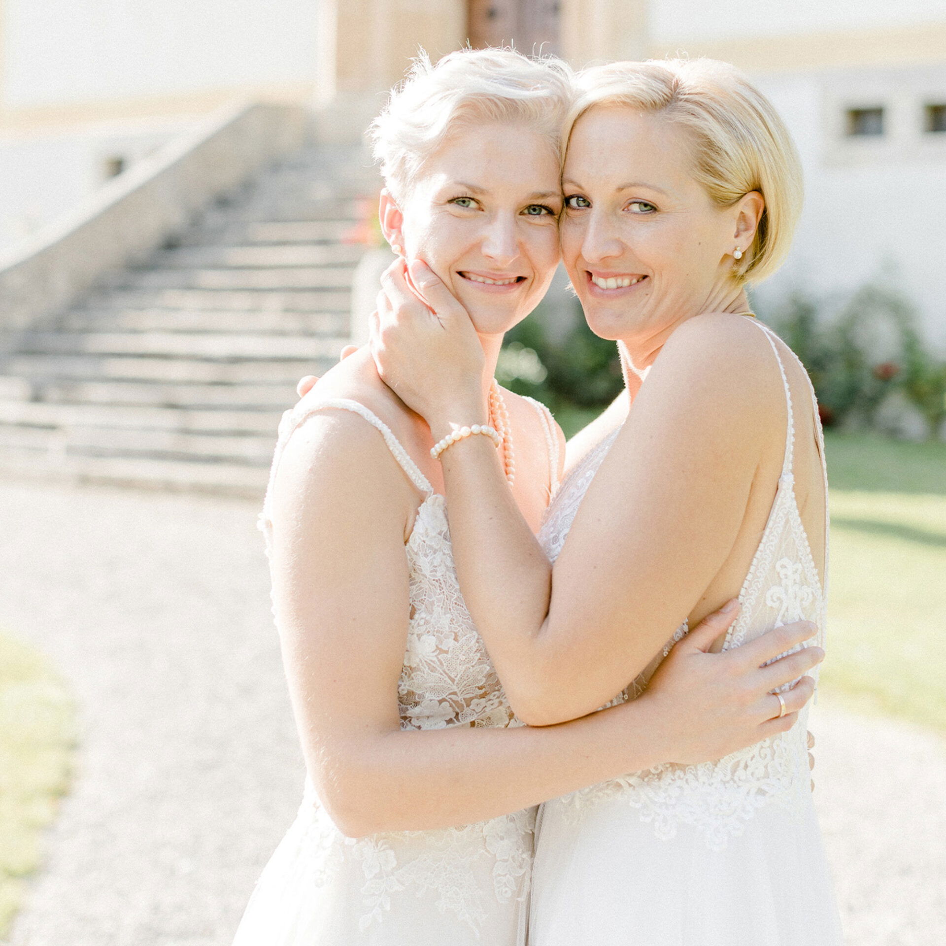 Ein fröhliches lesbisches Paar in eleganten Brautkleidern umarmt sich herzlich und genießt ihren besonderen Tag bei sonnigem Wetter im Freien.