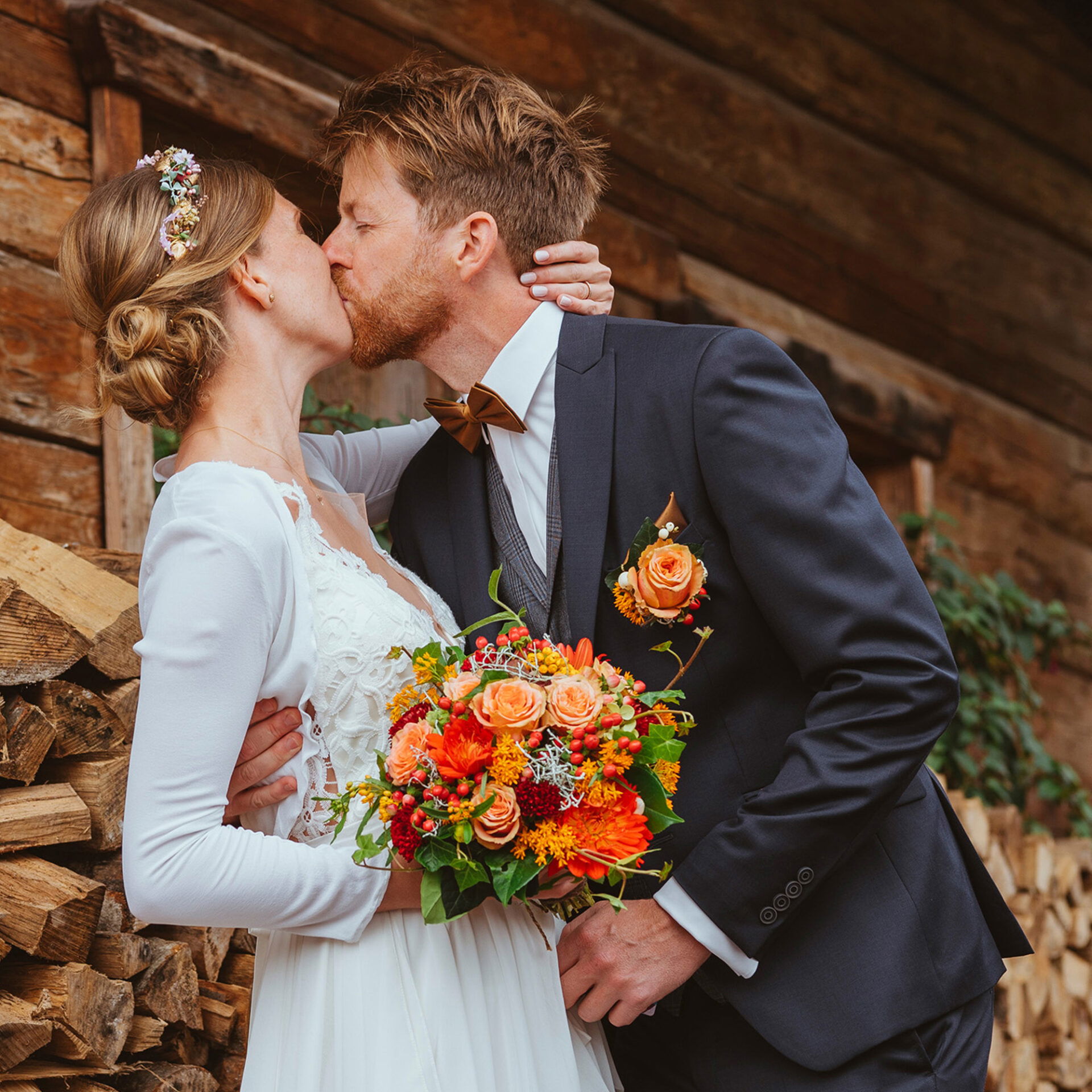 Das Brautpaar, in festlicher Kleidung, steht vor einer rustikalen Holzkulisse und teilt einen liebevollen Kuss. Die Braut trägt ein florales Diadem und hält einen leuchtend bunten Strauß, der Bräutigam trägt einen dunklen Anzug mit einem orangefarbenen Ansteckblümchen.