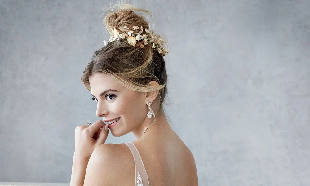 Nahaufnahme einer Braut mit luftigem Hochzeits-Dutt. Feine Blumen und goldene Blätter schmücken die Frisur, während dezente Ohrringe das Styling ergänzen. Eine wunderschöne Hochzeitsfrisur für romantische Bräute.
