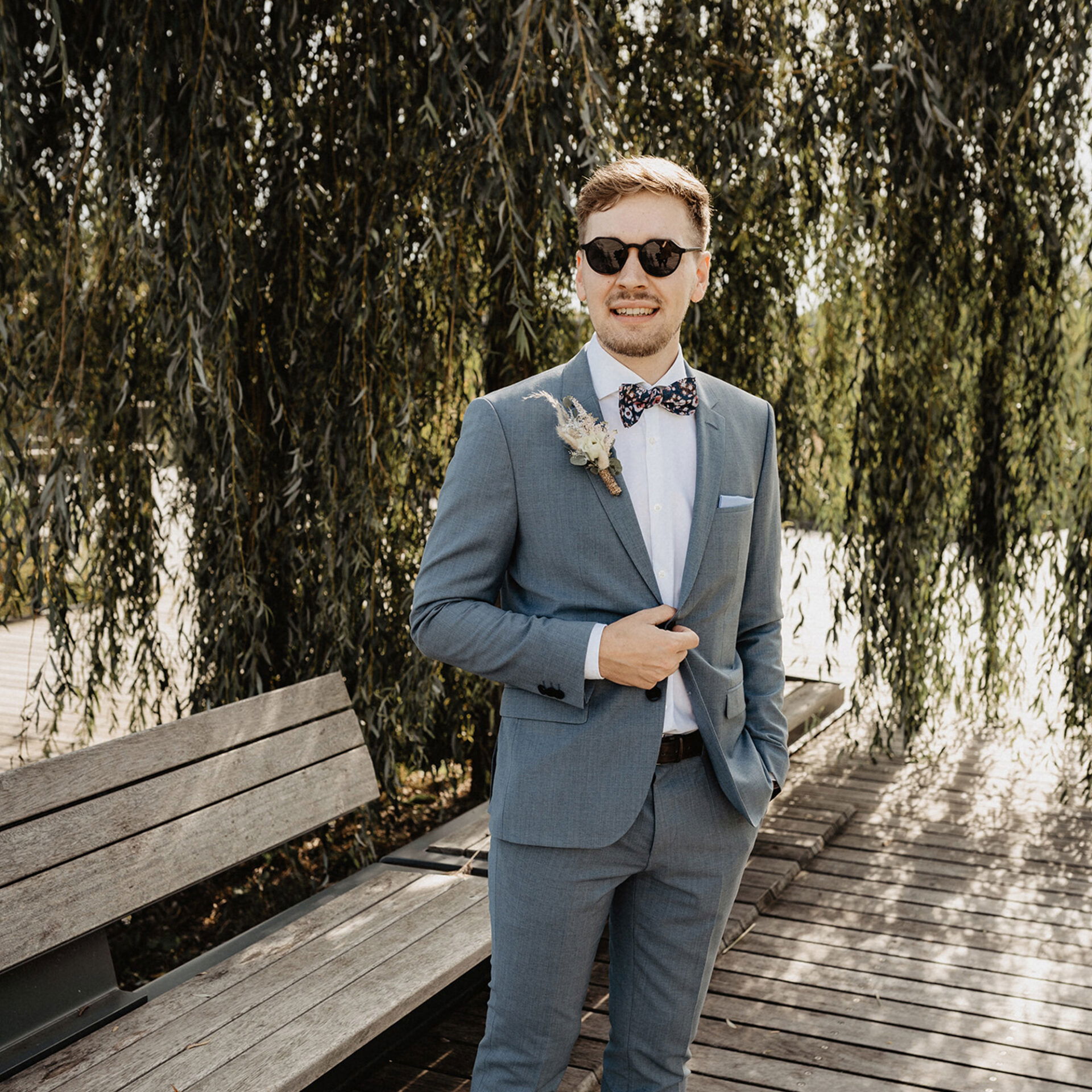 Der Bräutigam im grauen Anzug und mit einer farbenfrohen Fliege posiert auf einem Holzweg. Er trägt eine Sonnenbrille und steht vor einer Bank und einem Weidenbaum, was der Szenerie eine ruhige, natürliche Atmosphäre verleiht.