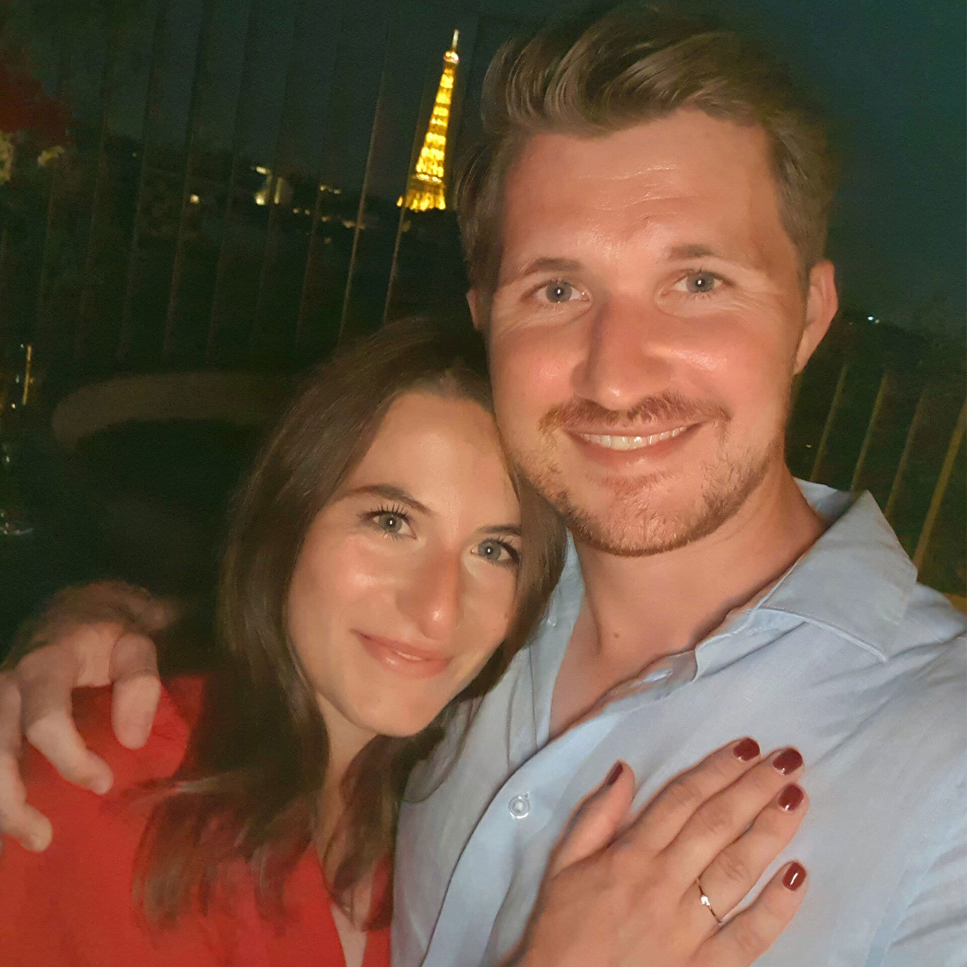Ein Paar posiert für ein Selfie bei Nacht in Paris mit dem beleuchteten Eiffelturm im Hintergrund. Die Frau trägt ein rotes Kleid und präsentiert ihren Verlobungsring, während sie den Arm um ihren Partner legt.