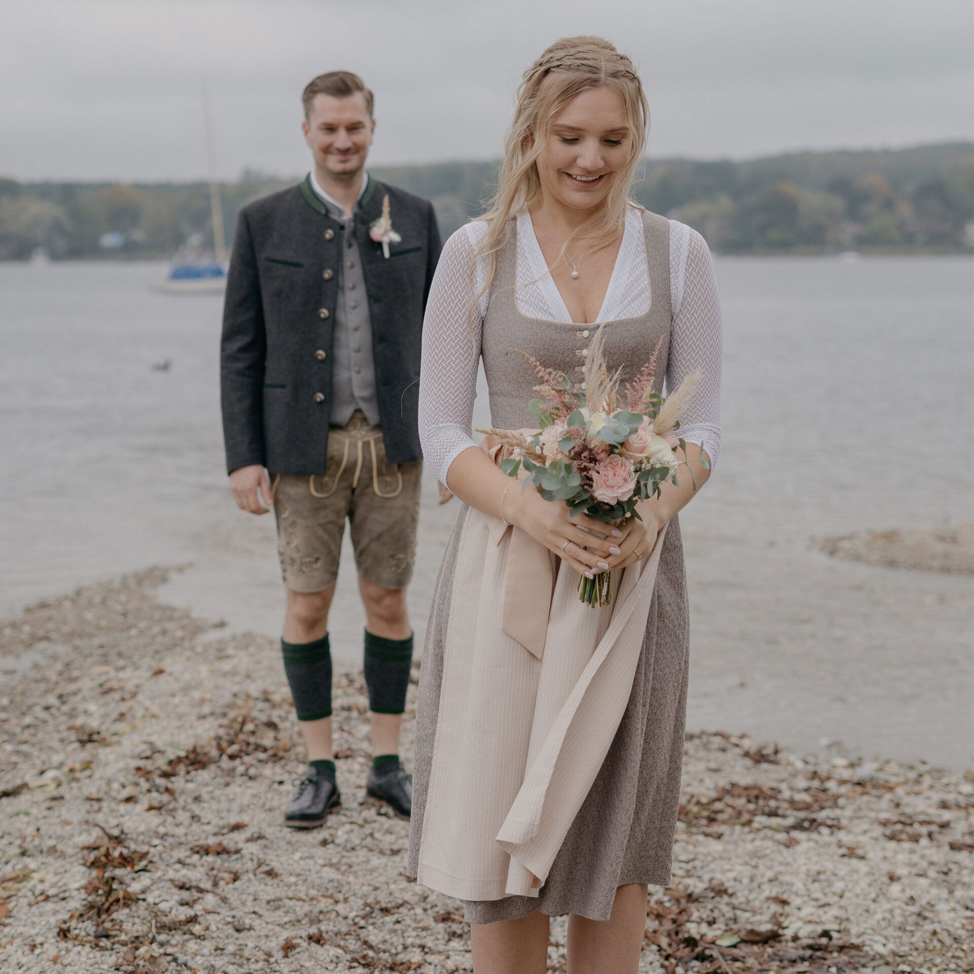 Die Braut in traditioneller Tracht hält einen zarten Blumenstrauß und lächelt verträumt, während sie am Kiesstrand steht. Der Bräutigam in bayerischer Tracht steht hinter ihr am See und beobachtet sie liebevoll.