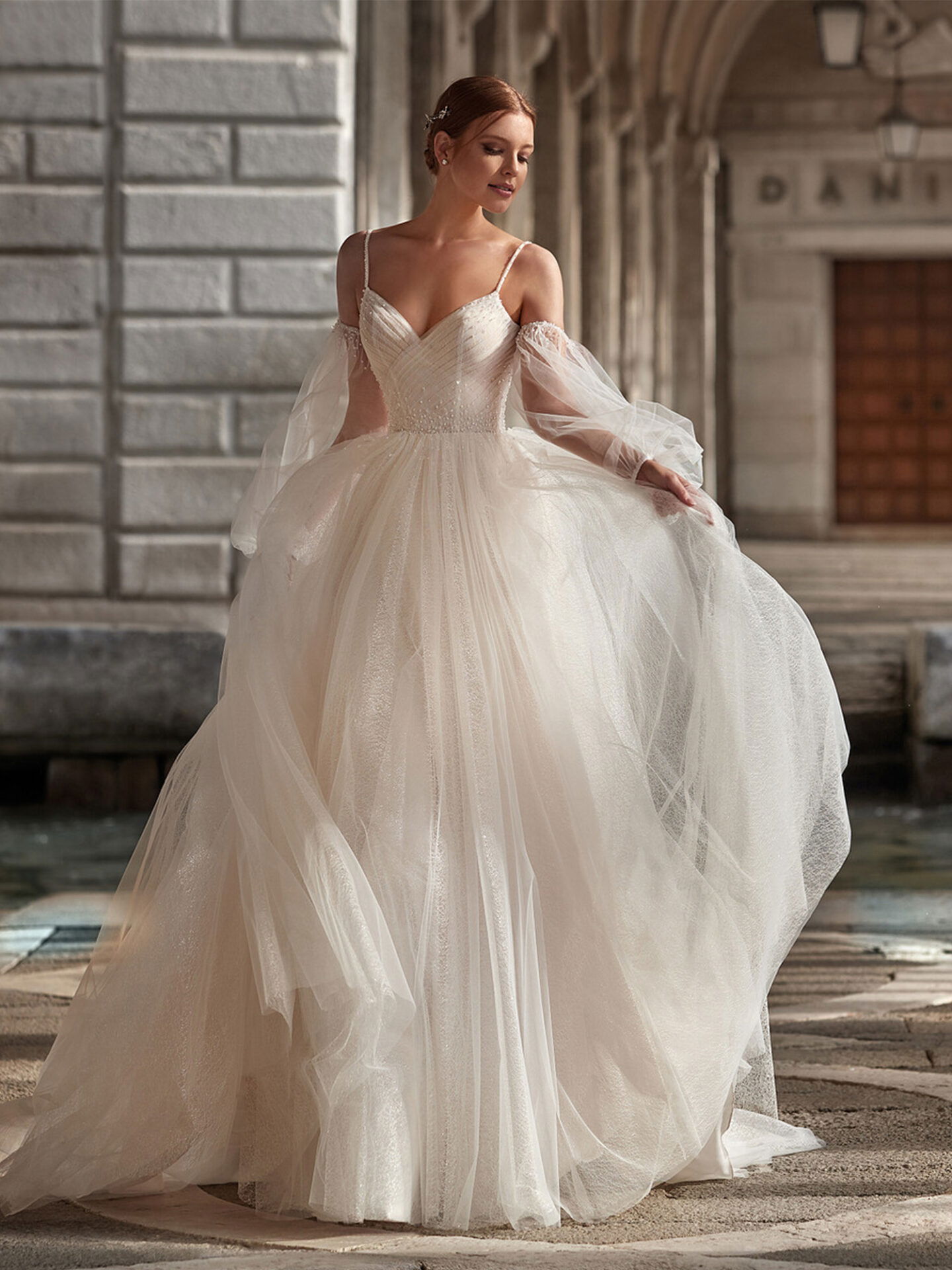 Eine Braut posiert in einem prächtigen, schulterfreien Brautkleid mit abnehmbaren Tüllärmeln vor einem historischen Gebäude. Der voluminöse Rock des Kleides bewegt sich leicht, während sie in eleganter Haltung dasteht.