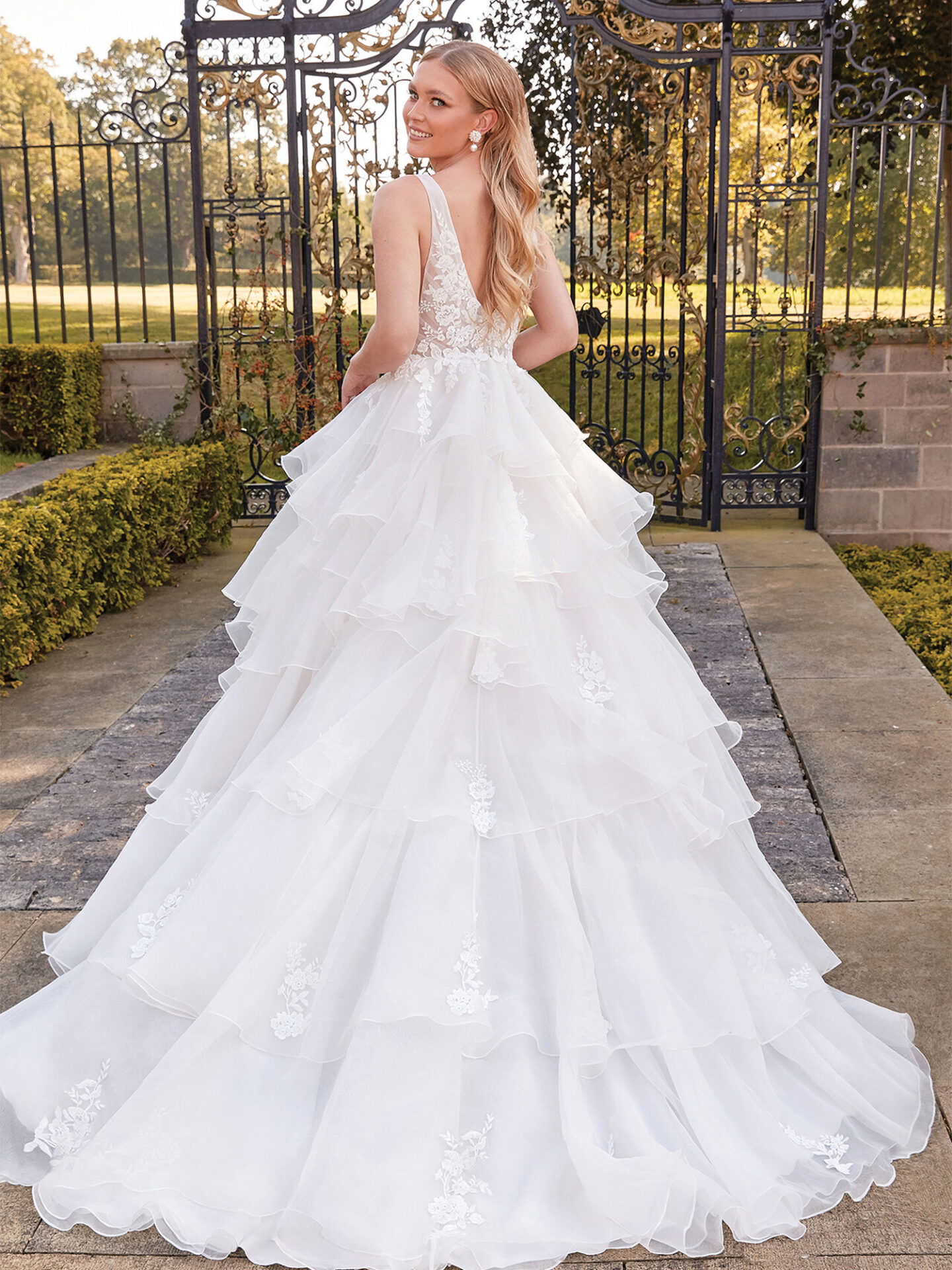 Eine Frau in einem eleganten Duchesse-Kleid mit einem tief ausgeschnittenen Rücken posiert vor einem Tor. Der Rock des Kleides ist mit floralen Applikationen verziert, und die Braut strahlt beim Blick über ihre Schulter.