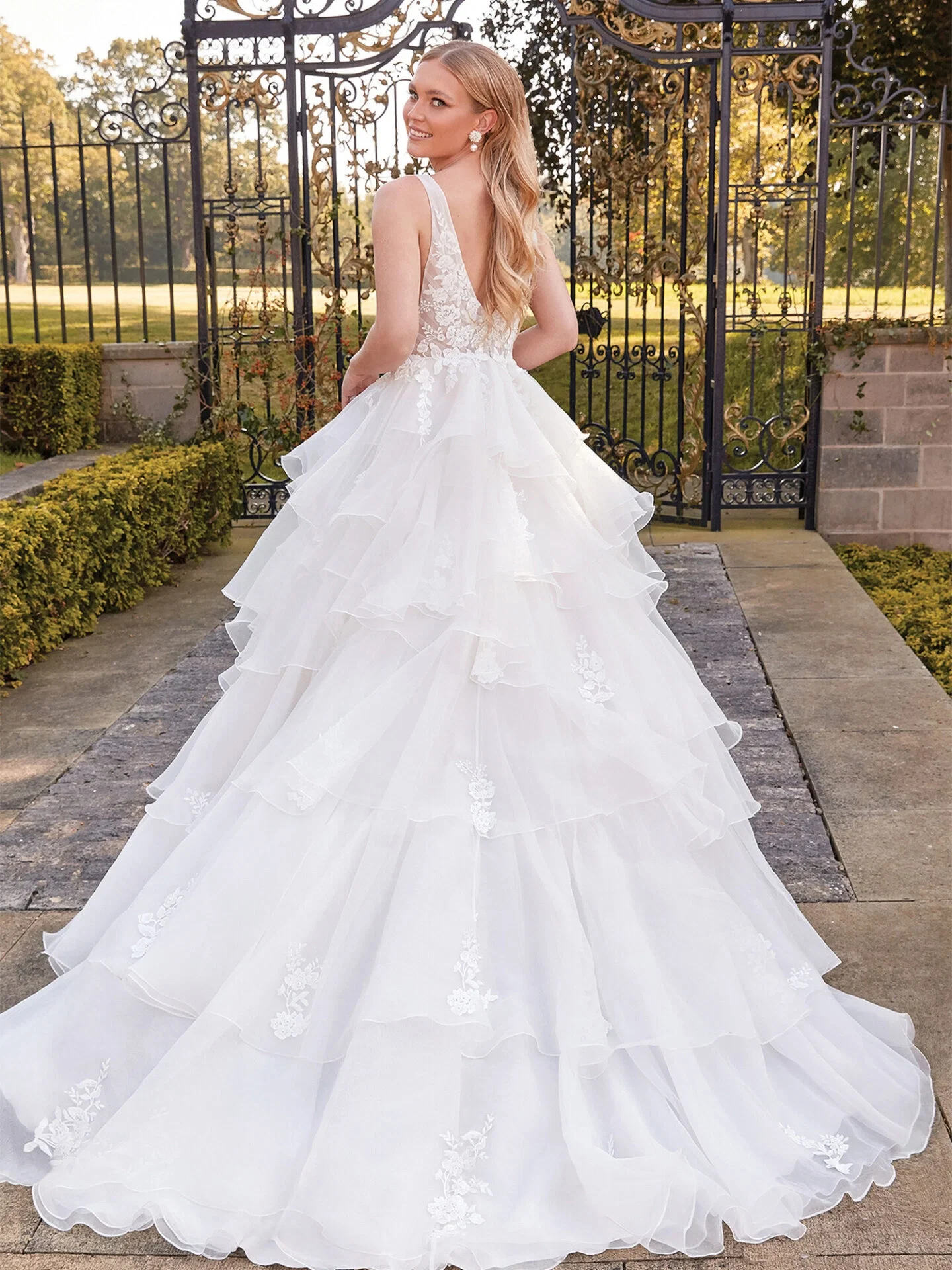 Eine Frau in einem eleganten Duchesse-Kleid mit einem tief ausgeschnittenen Rücken posiert vor einem Tor. Der Rock des Kleides ist mit floralen Applikationen verziert, und die Braut strahlt beim Blick über ihre Schulter.