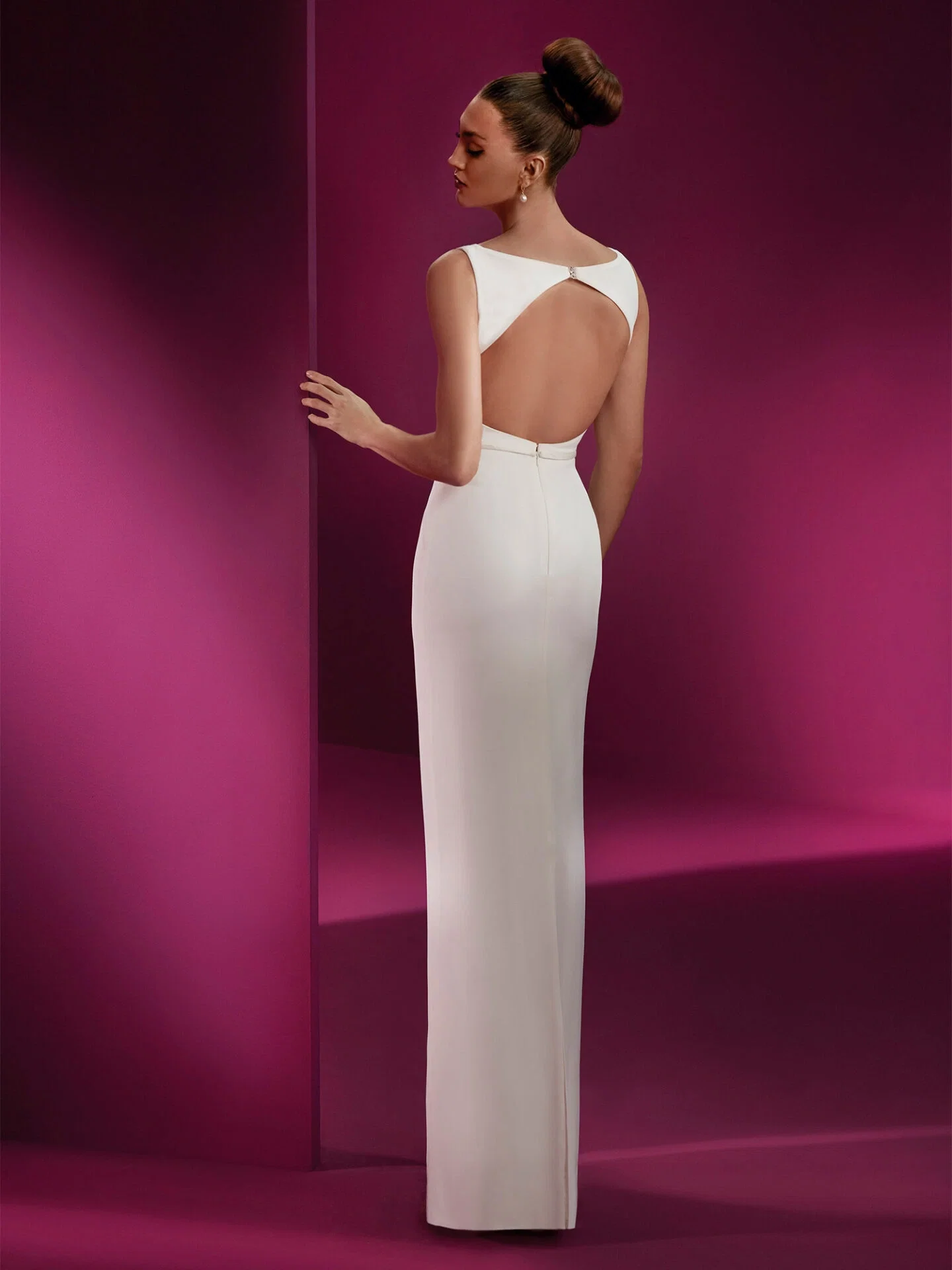 Eine Frau in einem eng anliegenden, rückenfreien weißen Etui-Kleid steht vor einer lilafarbenen Wand. Der schlichte Schnitt des Kleides kombiniert mit dem offenen Rücken verleiht dem Look eine zeitlose, raffinierte Eleganz.