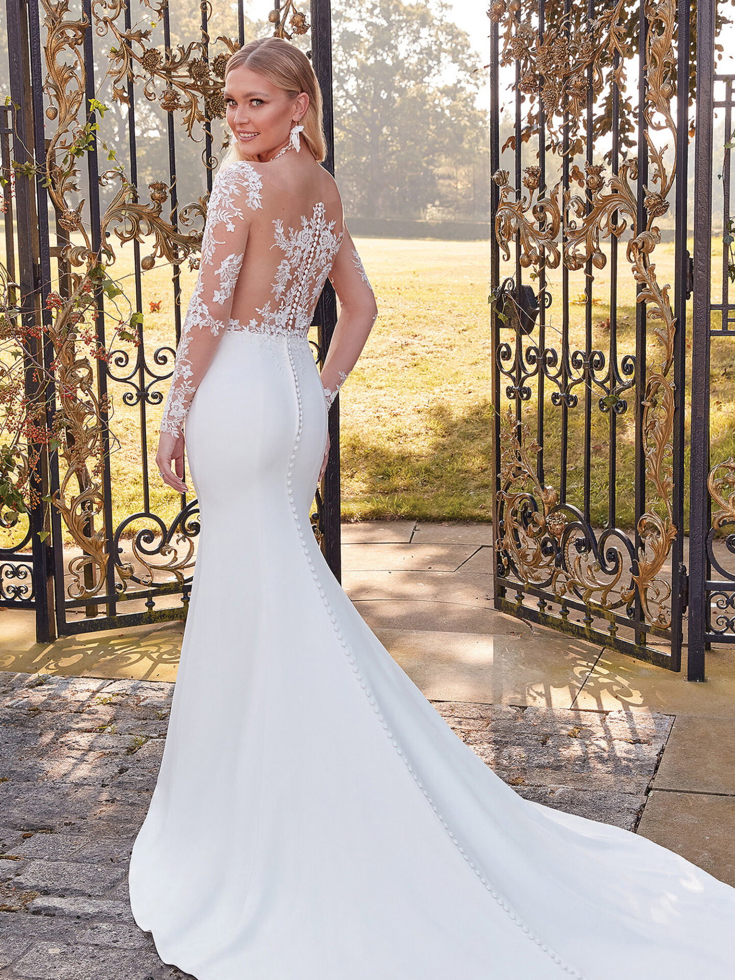 Eine Braut in einem eng anliegenden Brautkleid mit floraler Spitze und langen Ärmeln blickt lächelnd über die Schulter. Das Kleid betont ihre Figur und endet in einer langen Schleppe, während sie vor einem verzierten Tor posiert.