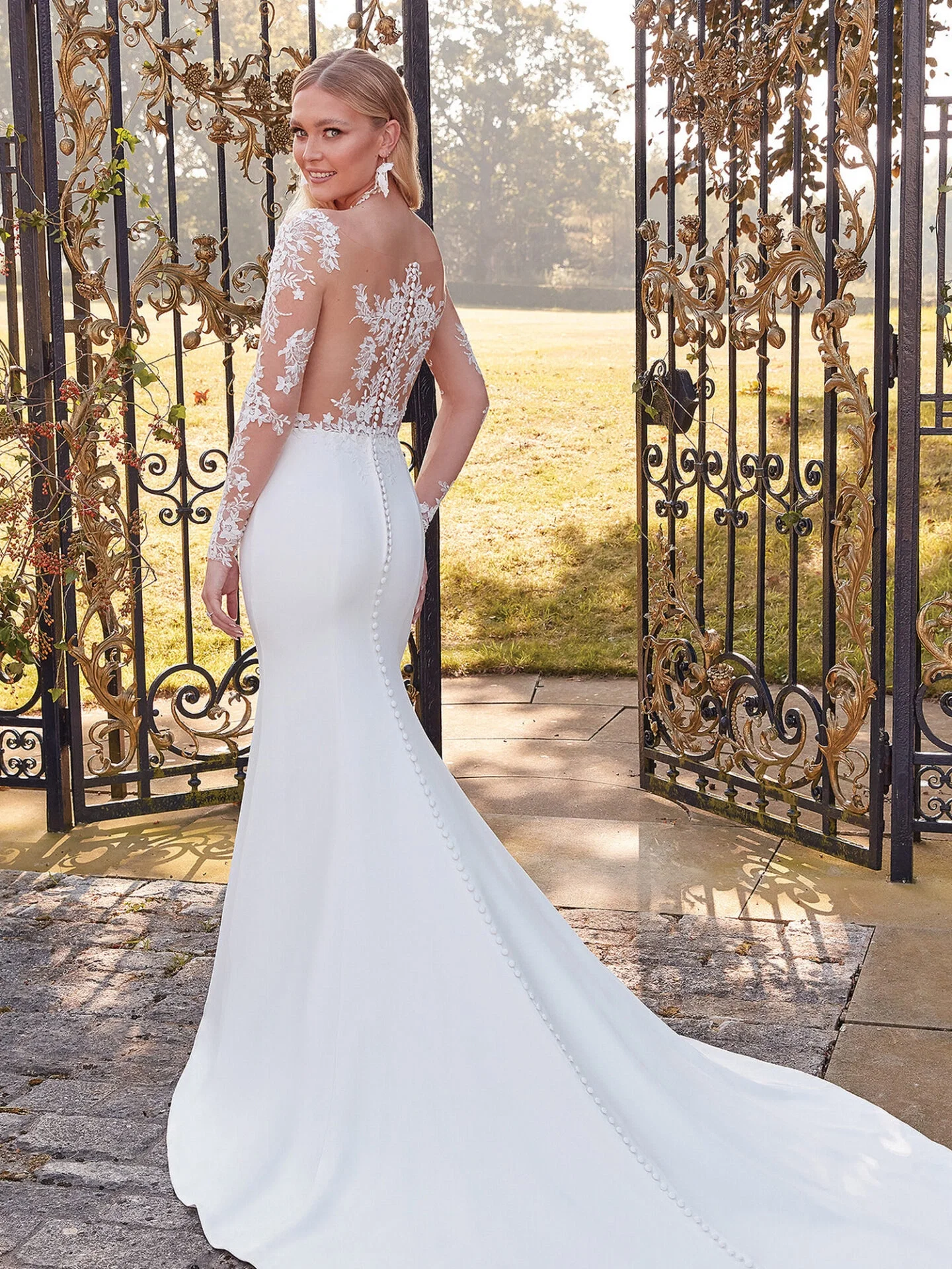 Eine Braut in einem eng anliegenden Brautkleid mit floraler Spitze und langen Ärmeln blickt lächelnd über die Schulter. Das Kleid betont ihre Figur und endet in einer langen Schleppe, während sie vor einem verzierten Tor posiert.