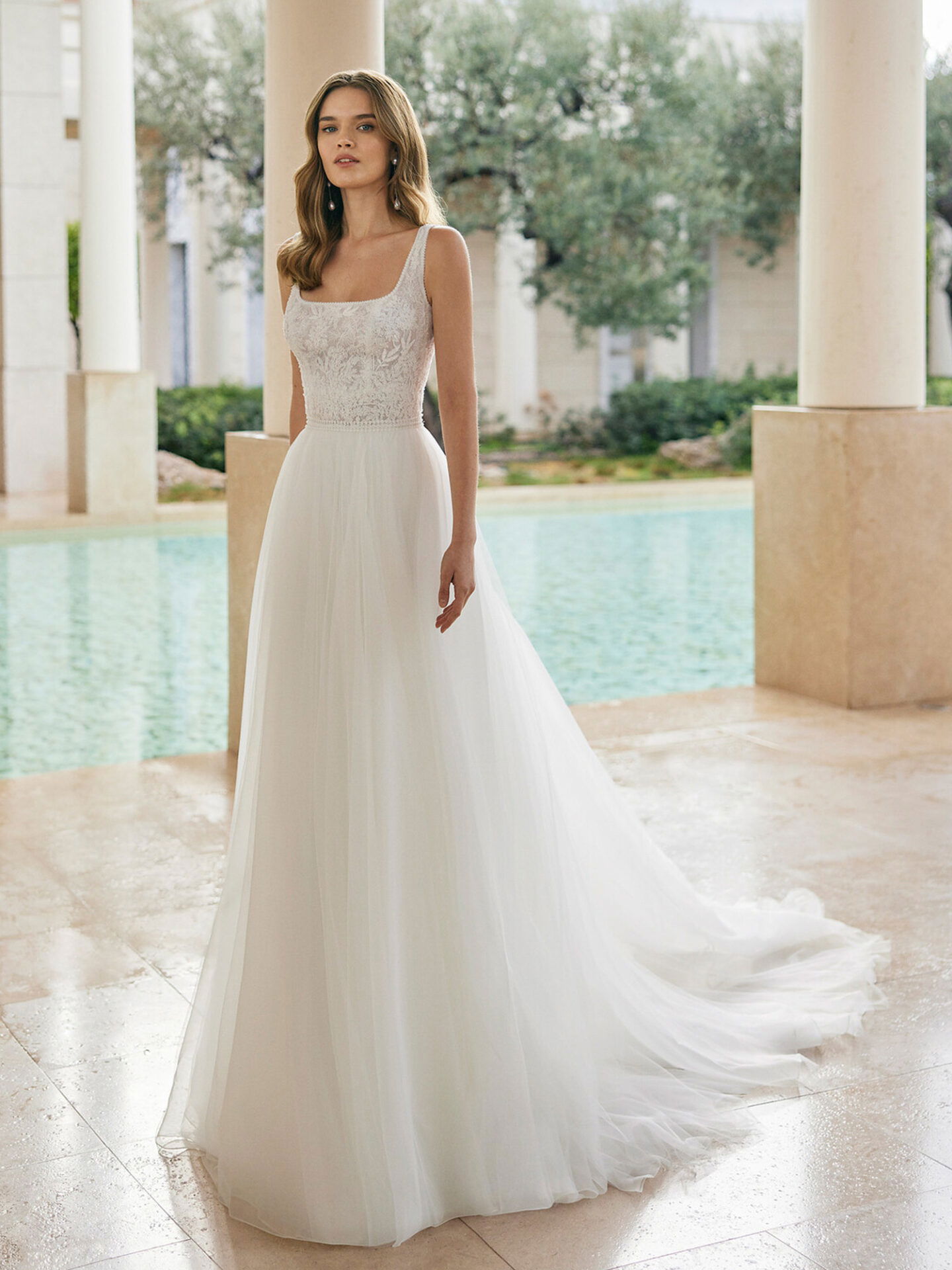 Eine Braut trägt ein minimalistisches, bodenlanges Kleid mit einem verzierten Mieder und breiten Trägern. Sie steht in einer eleganten Umgebung mit Blick auf einen Pool, während das weiche Licht den Stoff ihres Kleides zum Leuchten bringt.