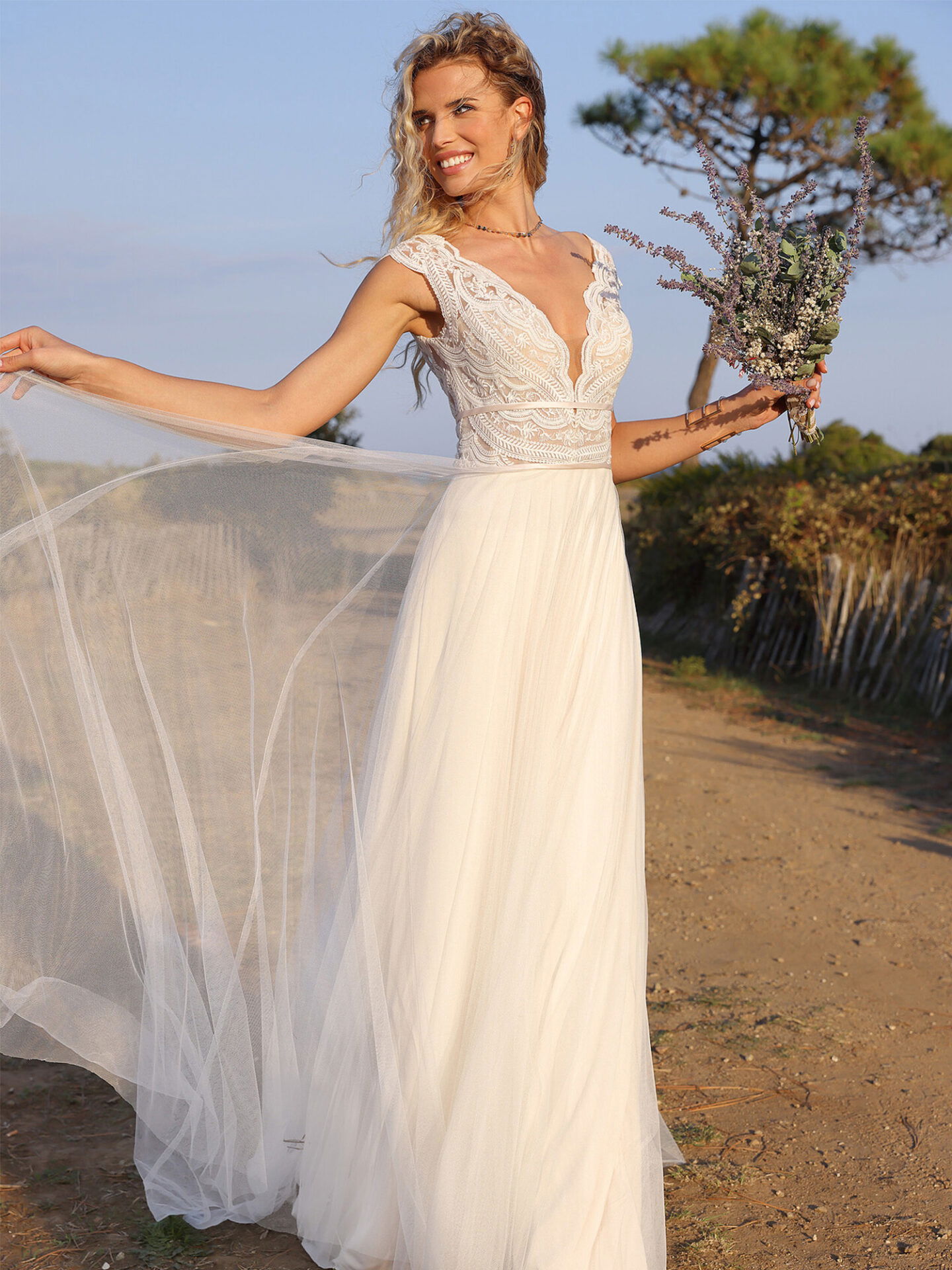 Eine Braut in einem leichten, fließenden I-Linien-Kleid mit einem tiefen V-Ausschnitt aus Spitze posiert draußen in einer sommerlichen Landschaft. Sie hält einen Lavendelstrauß und lächelt, während sie den Tüllrock ihres Kleides elegant in die Luft hebt.