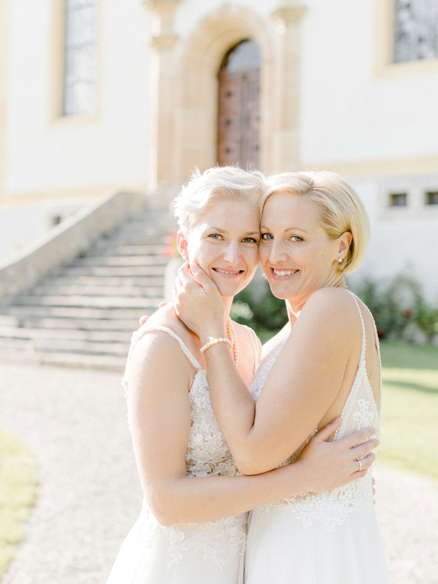 Dieses Bild zeigt zwei Bräute in weißen Kleidern, die sich eng umarmen und in die Kamera lächeln. Im Hintergrund sieht man ein altes Gebäude und eine Treppe, die den idyllischen Ort ihrer Hochzeit unterstreichen, während sie ihren besonderen Moment genießen.
