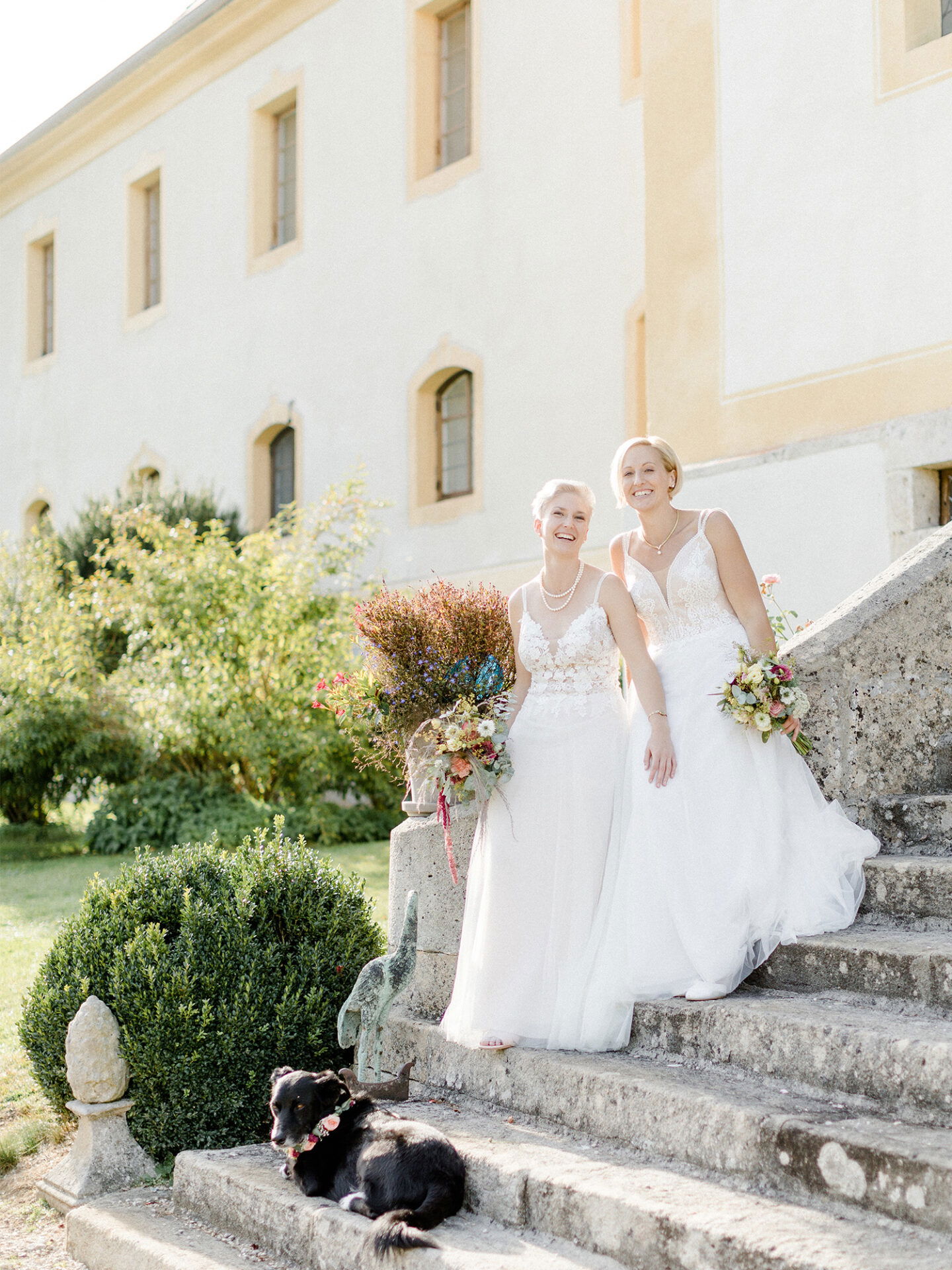Vor einem beeindruckenden historischen Gebäude posieren zwei Bräute in weißen Kleidern auf den Steinstufen. Ihre Blumensträuße und der zufriedene, liegende Hund mit Blumenschmuck an der Seite fangen eine idyllische Hochzeitsatmosphäre ein.