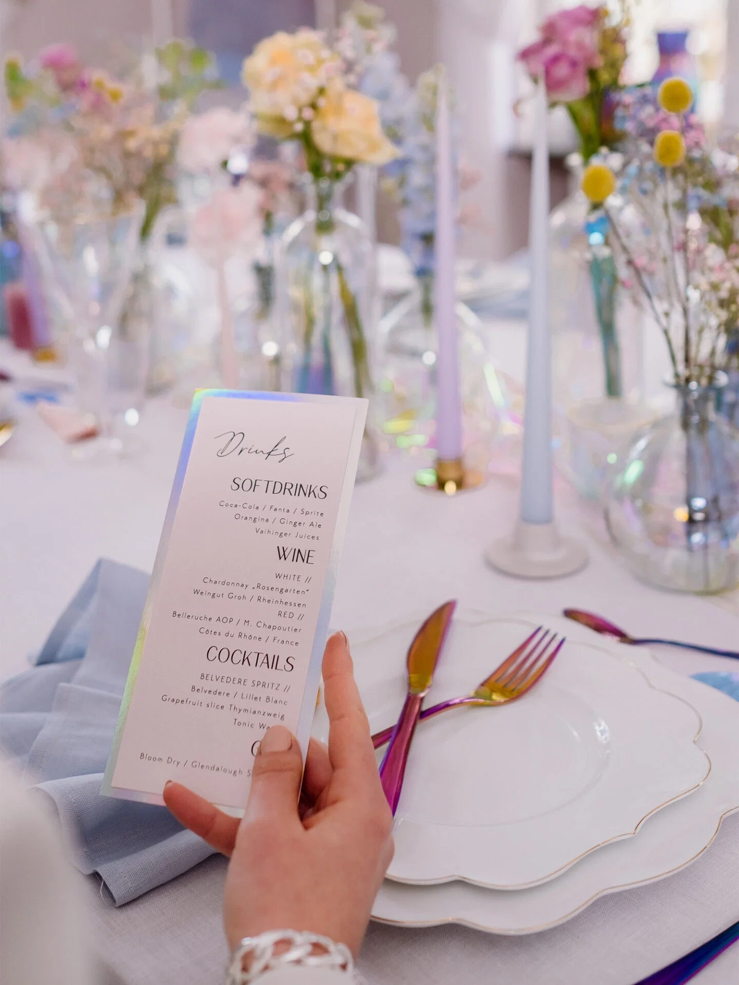Ein elegant gedeckter Tisch mit Blumenarrangements in Glasvasen und minimalistischen, pastellfarbenen Kerzen. Eine Person hält eine Menükarte mit den Getränken, und das Regenbogenbesteck sowie die mit Gold verzierte Teller setzen stilvolle Akzente.