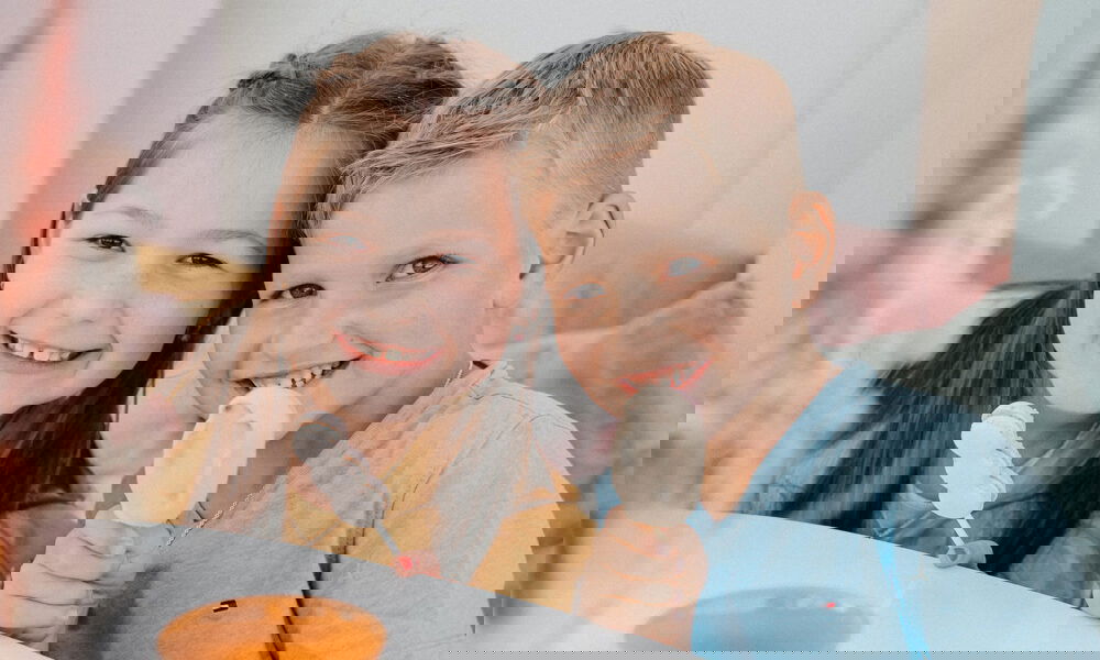 Zwei fröhliche Kinder sitzen gemeinsam am Tisch und naschen Süßigkeiten, die Szene wirkt hell und freundlich.