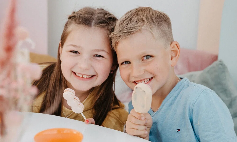 Zwei fröhliche Kinder sitzen gemeinsam am Tisch und naschen Süßigkeiten, die Szene wirkt hell und freundlich.