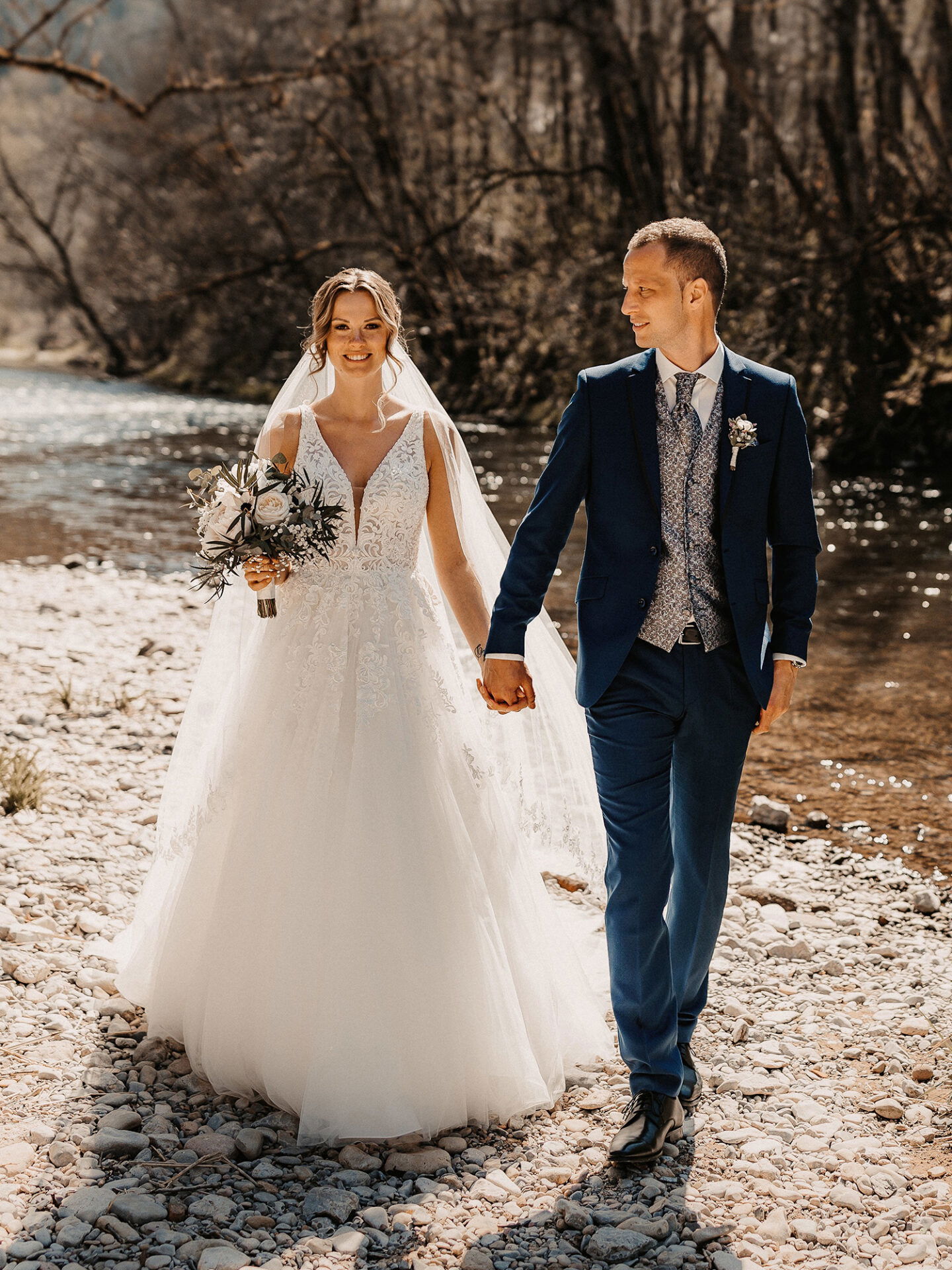 In einem idyllischen Setting am Fluss, hält die Braut mit ihrem fließenden weißen Kleid den Bräutigam bei der Hand, beide sehen entspannt und glücklich aus.