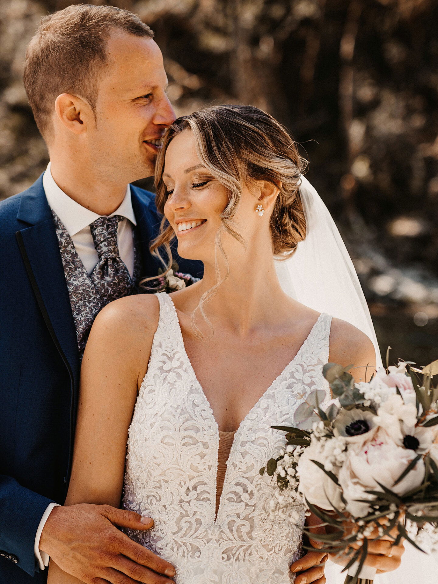 Romantisches Portrait eines Paares in Hochzeitskleidung: Die Braut in einem filigranen Kleid mit tiefem Ausschnitt, der Bräutigam hält sie fest im Arm und beide lächeln zufrieden.
