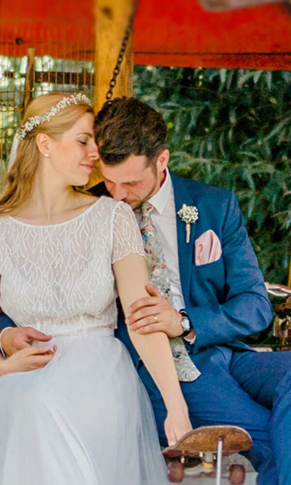 Ein romantischer Moment zwischen Braut und Bräutigam auf einem Karussell. Die Braut, mit einem funkelnden Diadem und einem fließenden Brautkleid, genießt die Umarmung des Bräutigams, der in einem schicken blauen Anzug mit gemusterter Krawatte gekleidet ist. Die Szene strahlt Ruhe und Nähe aus.