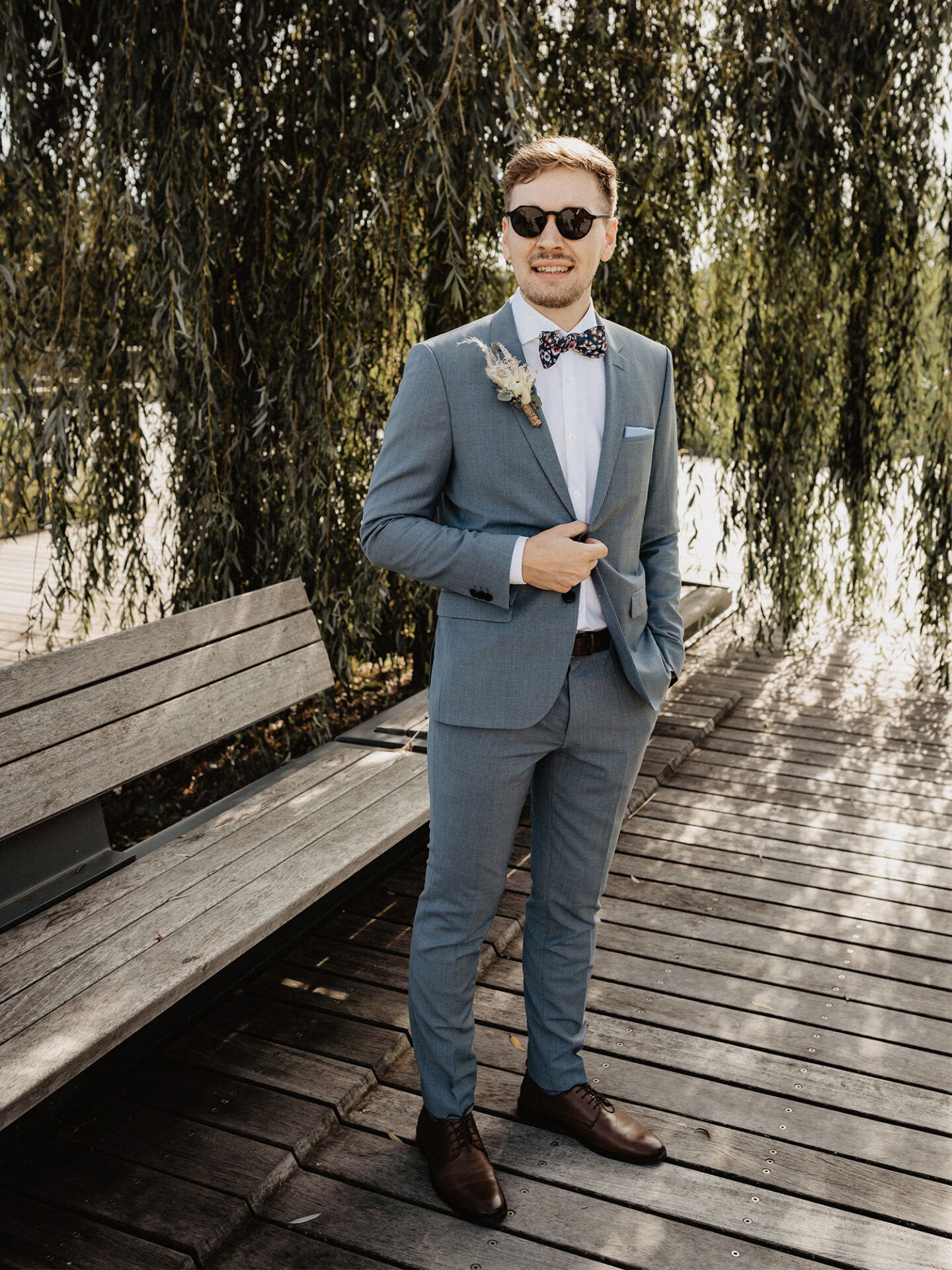 Ein stilvoll gekleideter Mann mit Anzug und Sonnenbrille steht auf einem hölzernen Steg vor einem Weidenbaum. Seine Pose ist lässig, und das natürliche Licht unterstreicht die entspannte Stimmung.