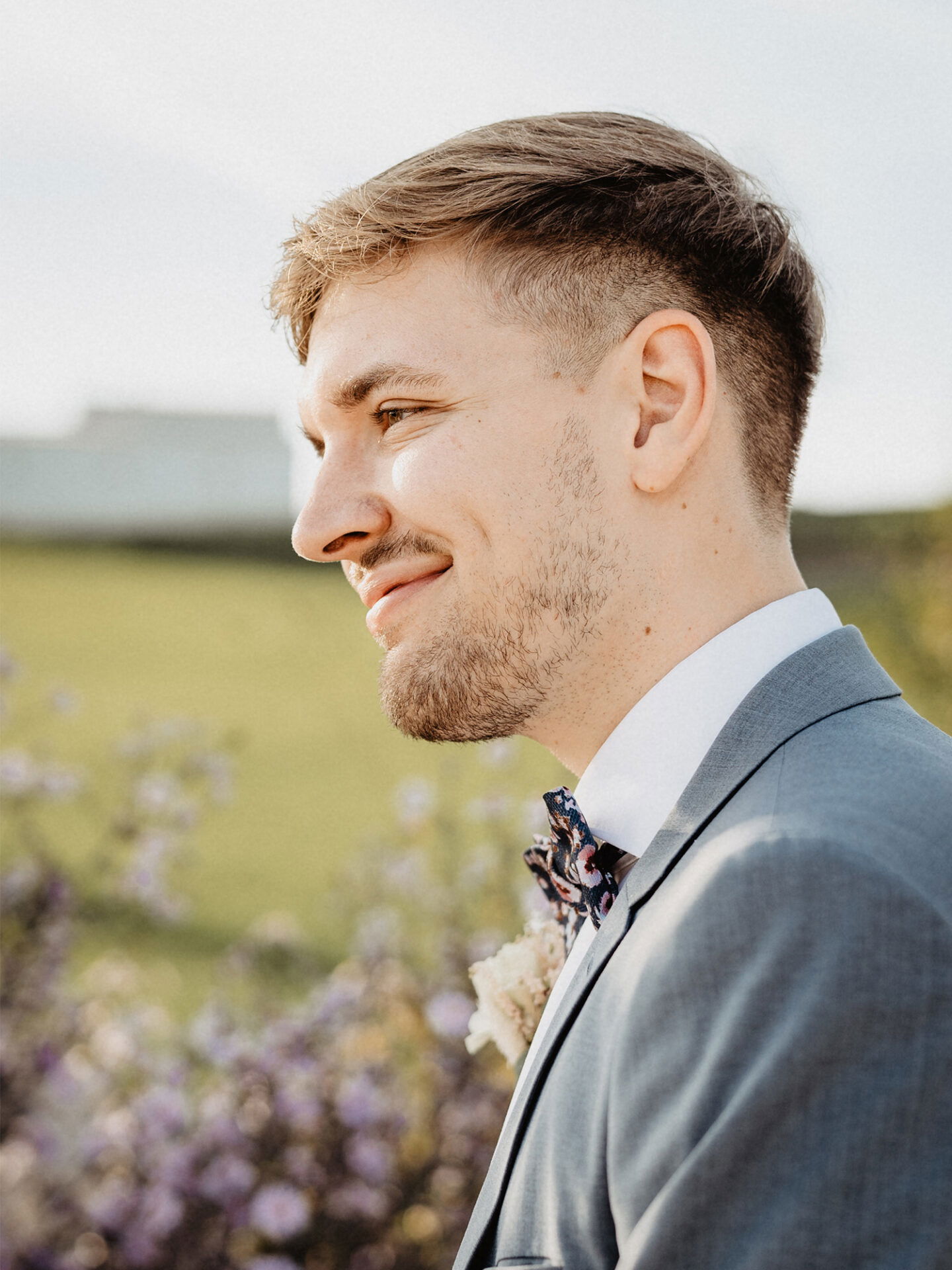 Ein Bräutigam im grauen Anzug mit gemusterter Fliege lächelt in die Ferne. Im Hintergrund sieht man blühende Blumen und eine ländliche Szenerie.