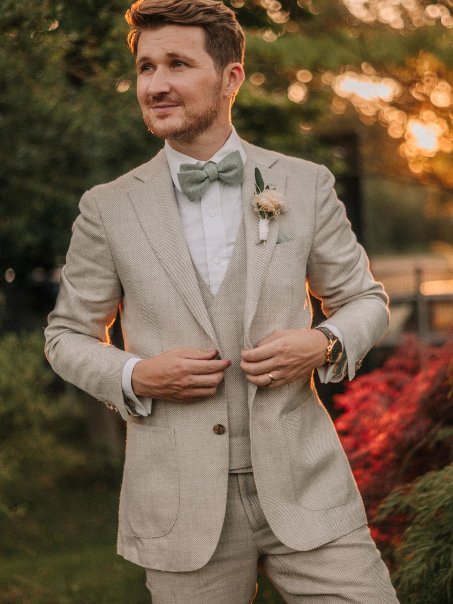 Ein eleganter Bräutigam in einem hellen Anzug und grünem Accessoire posiert bei Sonnenuntergang in einem Garten. Der Anzug und die Kulisse betonen den feierlichen Moment.