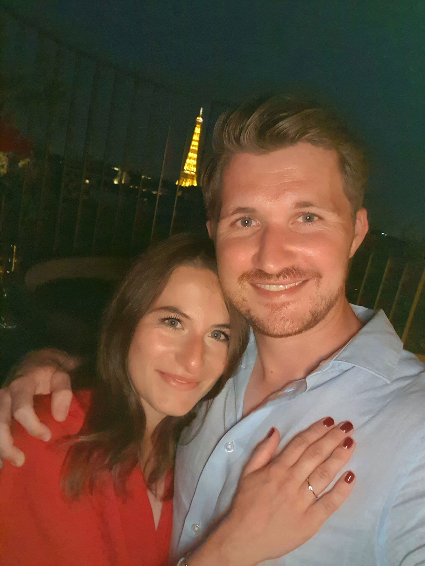 Ein Paar feiert ihre Verlobung in Paris und hält ein Selfie vor dem beleuchteten Eiffelturm. Die Frau trägt einen Verlobungsring und lächelt glücklich in die Kamera.