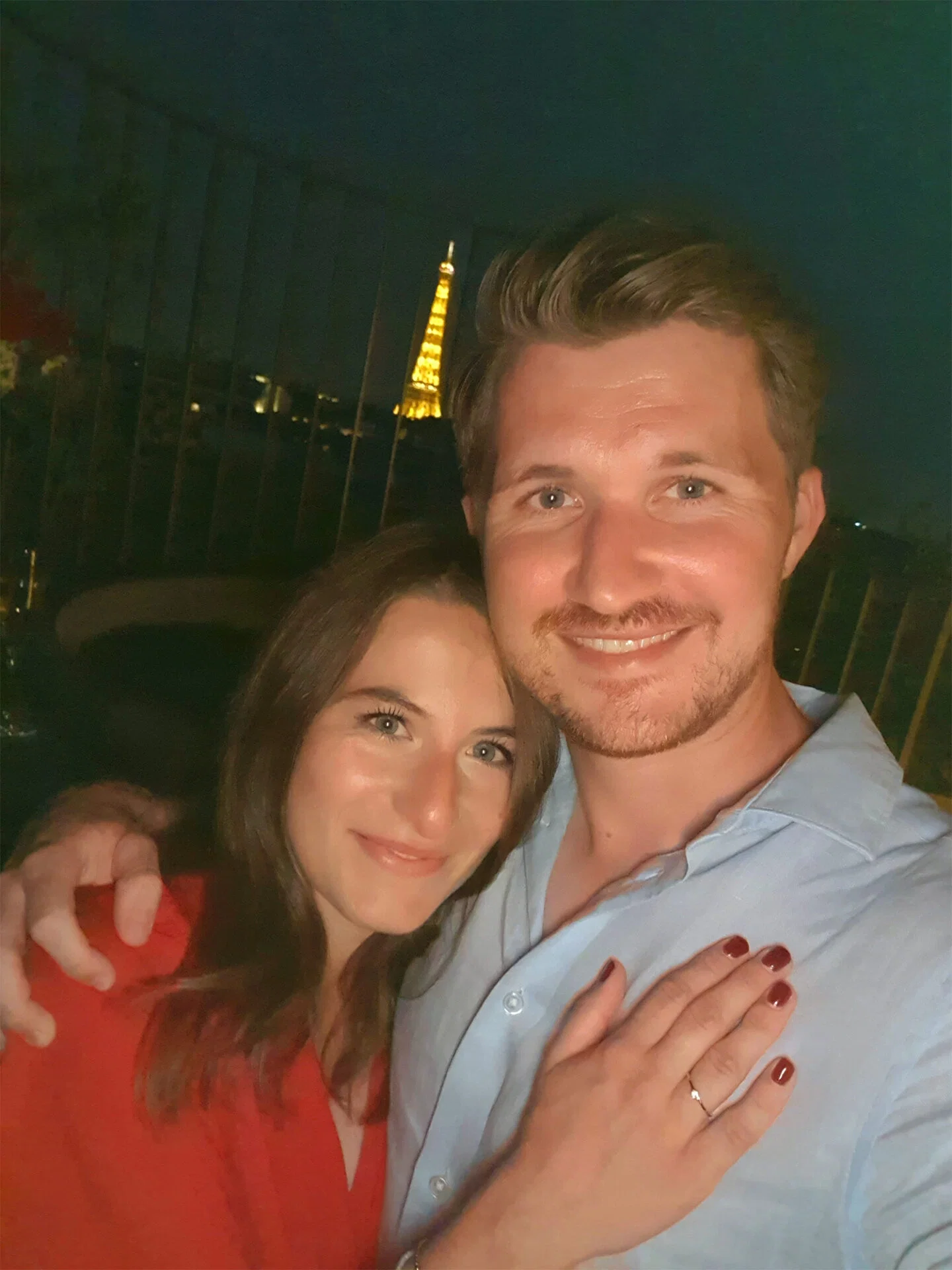 Ein Paar feiert ihre Verlobung in Paris und hält ein Selfie vor dem beleuchteten Eiffelturm. Die Frau trägt einen Verlobungsring und lächelt glücklich in die Kamera.