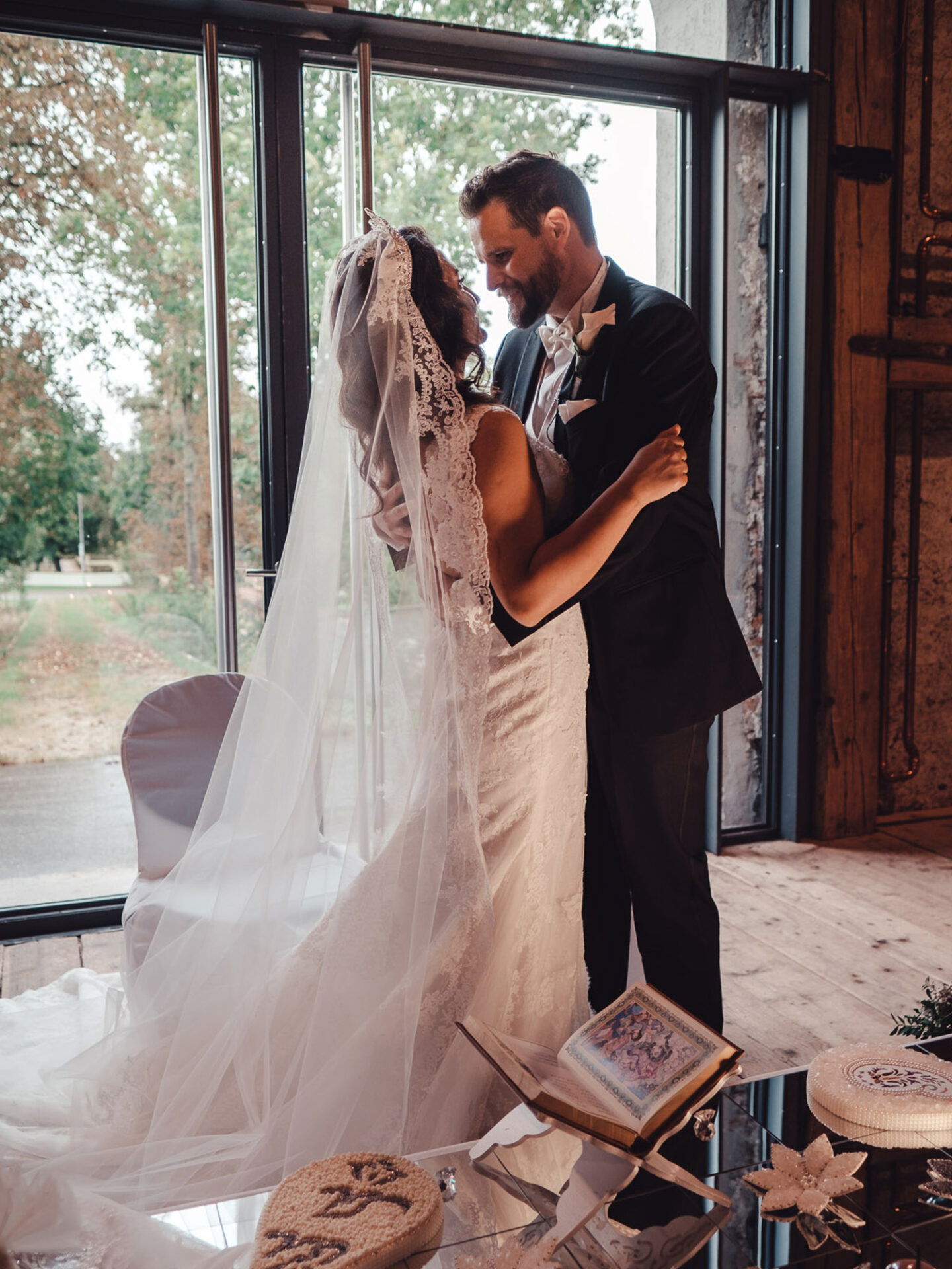 Ein frisch verheiratetes Paar steht in einer romantischen Umarmung vor einem raumhohen Fenster mit Blick auf die Natur. Die Braut trägt ein weißes Spitzenkleid und einen langen Schleier, der Bräutigam ist in Schwarz gekleidet. Im Vordergrund sind ein religiöses Buch und kunstvolle Dekorationen auf einem Glastisch zu sehen.
