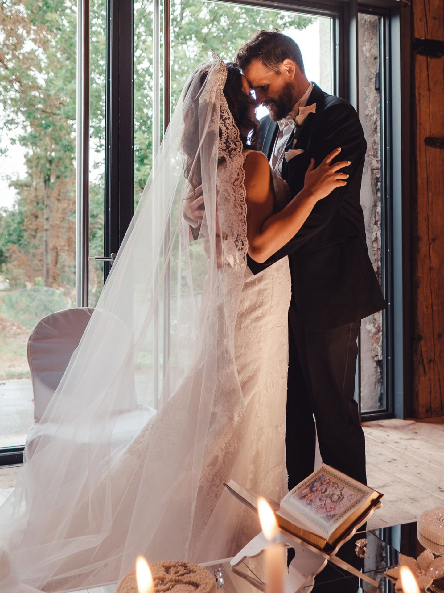 Ein verliebtes Brautpaar teilt einen intimen Moment in einem rustikalen Raum mit großen Fenstern. Die Braut trägt ein traditionelles Hochzeitskleid mit Spitze und Schleier, während der Bräutigam in einem formellen schwarzen Anzug zu sehen ist. Vor ihnen steht ein Tisch mit brennenden Kerzen und einem aufgeschlagenen Buch.