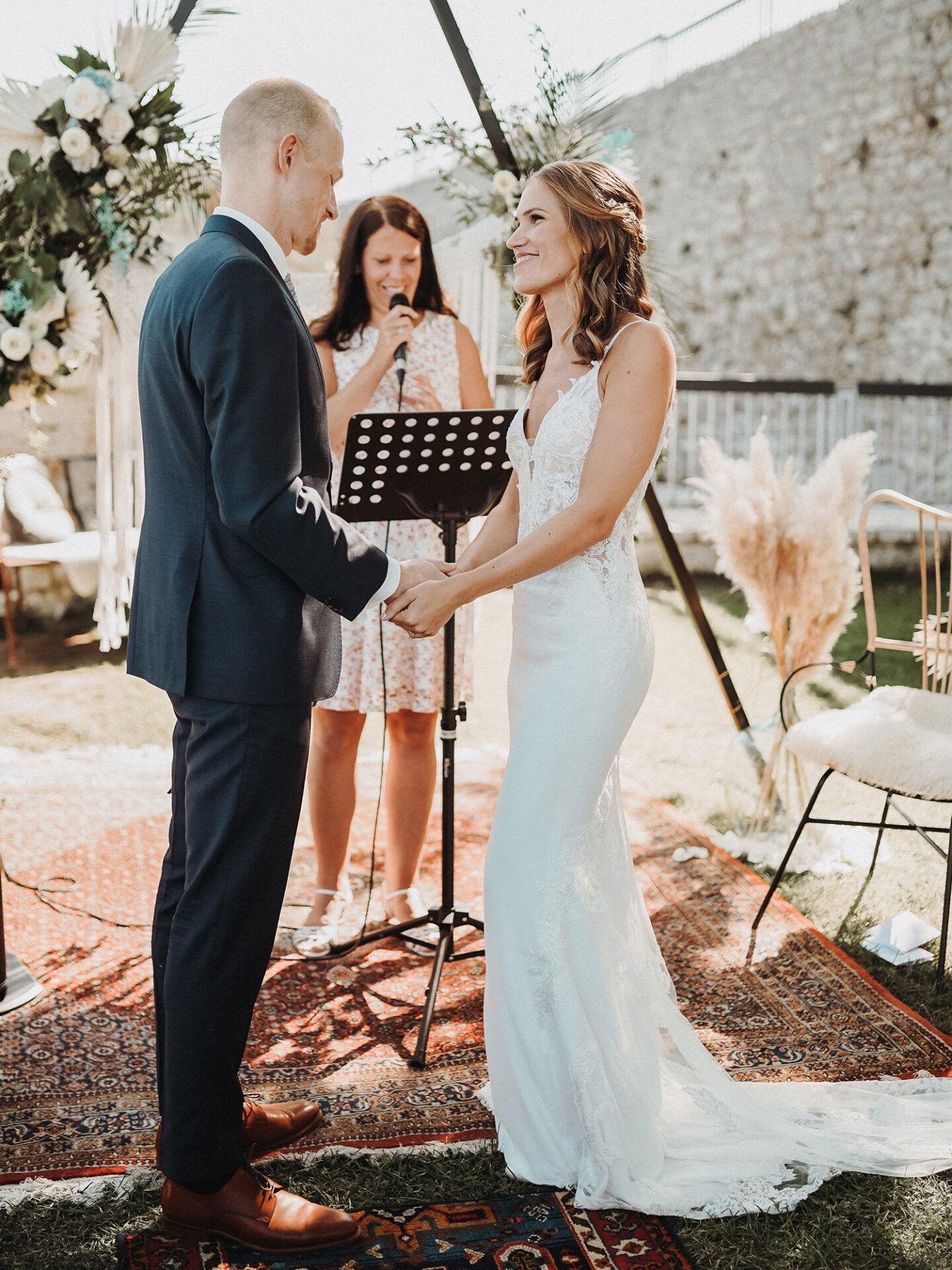 Ein glückliches Brautpaar steht sich gegenüber und hält sich an den Händen, während sie ihre Gelübde austauschen. Die Braut trägt ein eng anliegendes Spitzenkleid und der Bräutigam trägt einen formellen, dunklen Anzug. Die Zeremonie findet im Freien statt, und eine Rednerin spricht im Hintergrund.