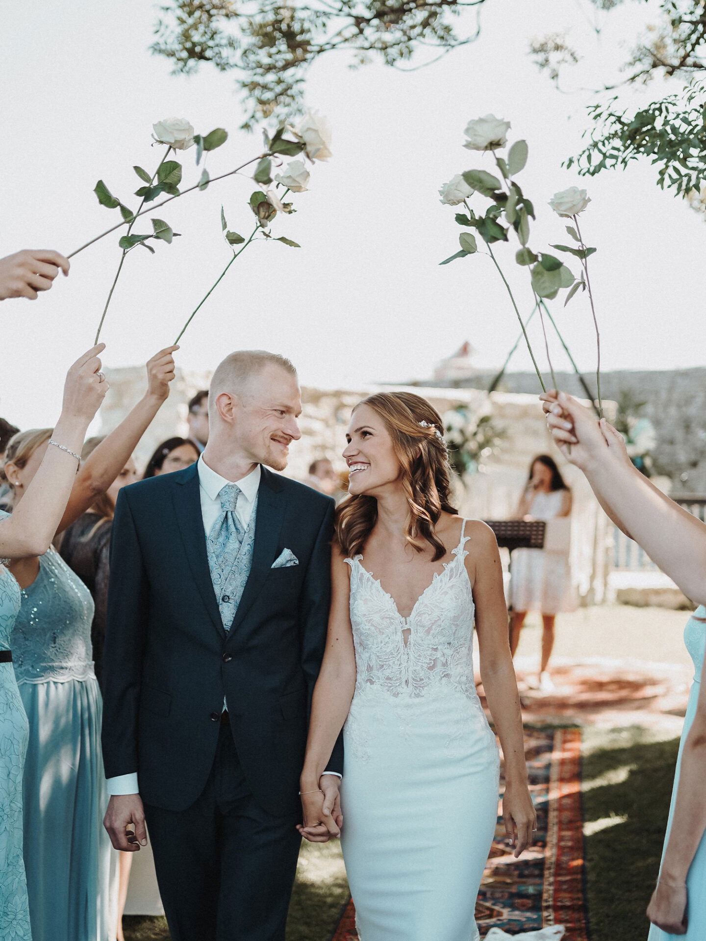 Ein frisch vermähltes Paar läuft Hand in Hand unter einem Spalier aus Rosen hindurch, die von den Gästen hochgehalten werden. Die Braut trägt ein verziertes Kleid aus Spitze und der Bräutigam einen stilvollen blauen Anzug. Beide lächeln glücklich und strahlen vor Freude.