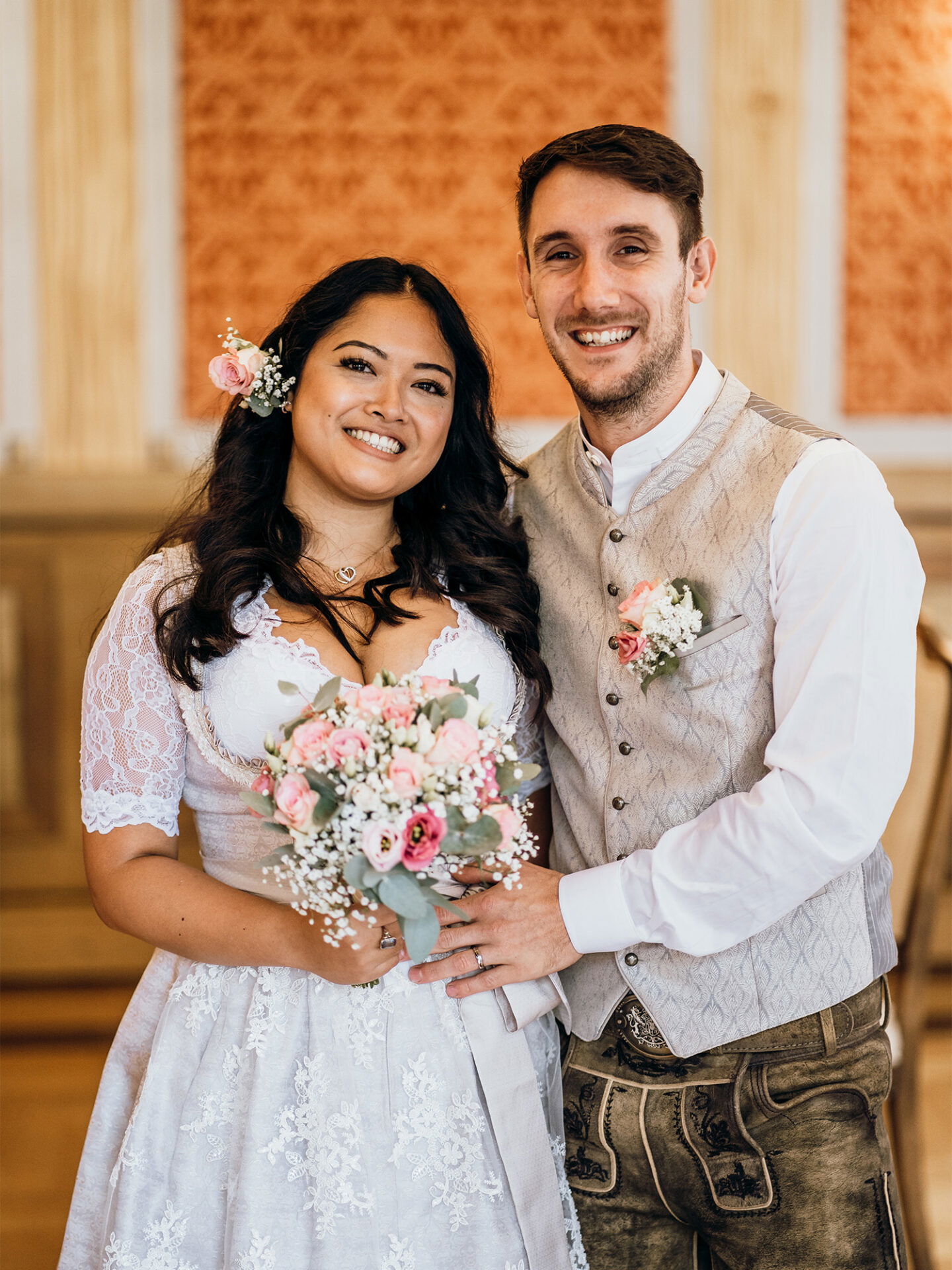 Ein glückliches Brautpaar steht Arm in Arm und lächelt. Die Braut trägt ein weißes, mit Blumen verziertes Kleid, und der Bräutigam ist in eine traditionelle bayerische Lederhose und Weste gekleidet. Die Braut hält einen rosafarbenen Blumenstrauß.