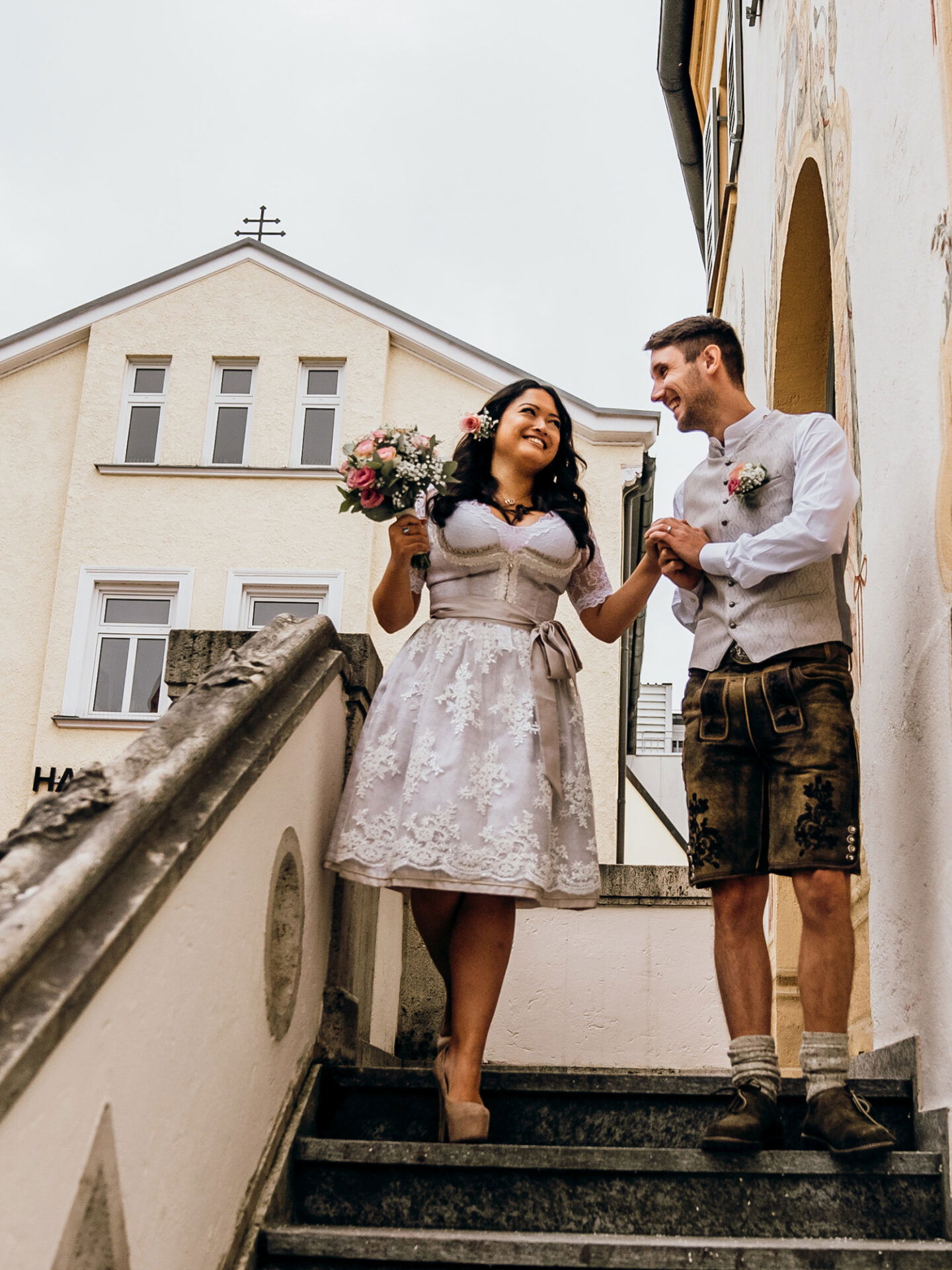 Ein frisch verheiratetes Paar läuft gemeinsam die Stufen hinunter. Die Braut trägt ein mit Blumen besticktes Dirndl und hält einen Blumenstrauß, während der Bräutigam in Lederhosen sie zärtlich an der Hand führt. Sie lächeln sich gegenseitig an.