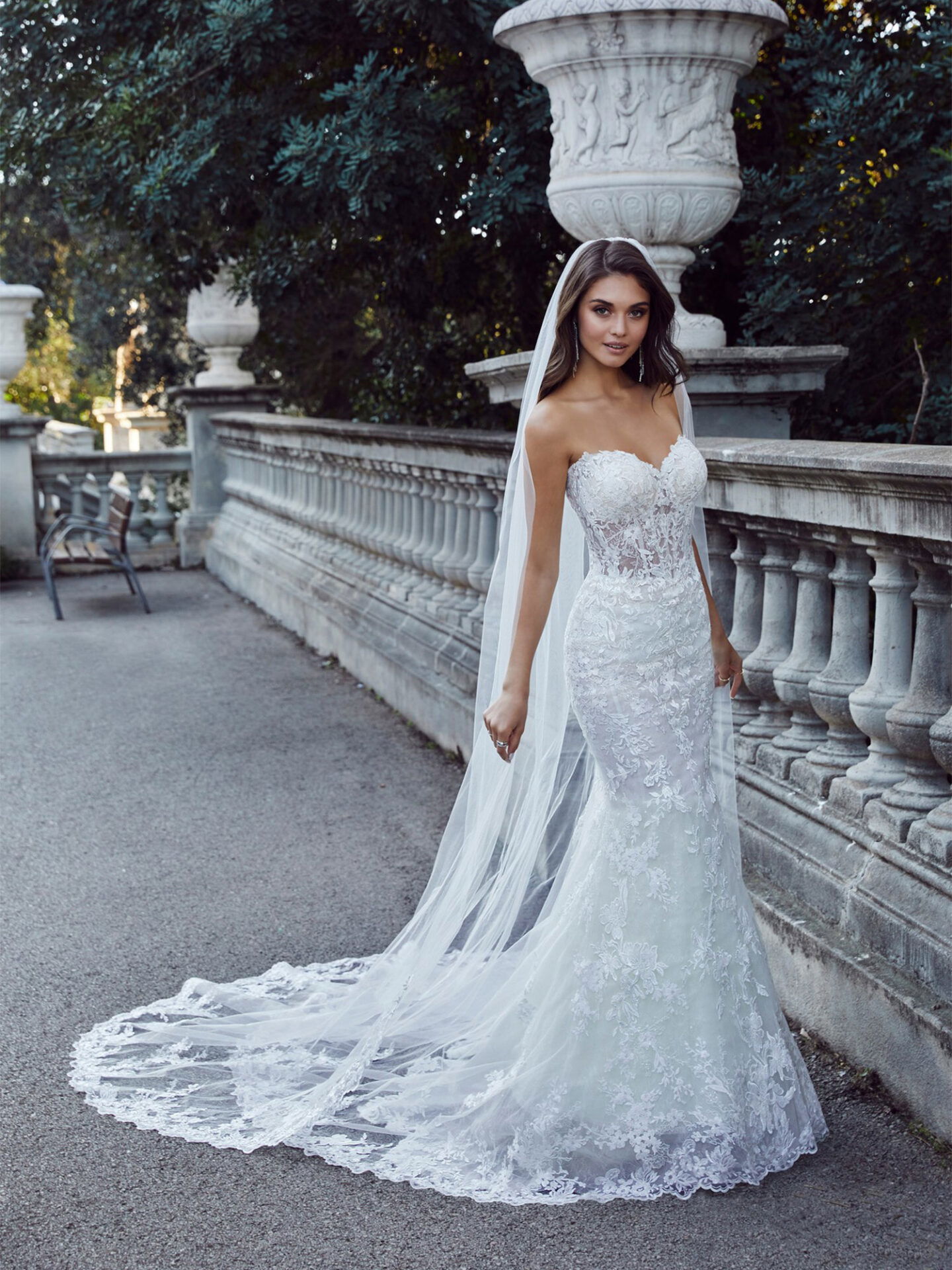 Eine Braut in einem schulterfreien, figurbetonten Mermaid-Kleid mit einer floralen Spitze und einer langen Schleppe steht vor einem historischen Bauwerk. Der Schleier fließt hinter ihr, während sie elegant in die Kamera blickt.
