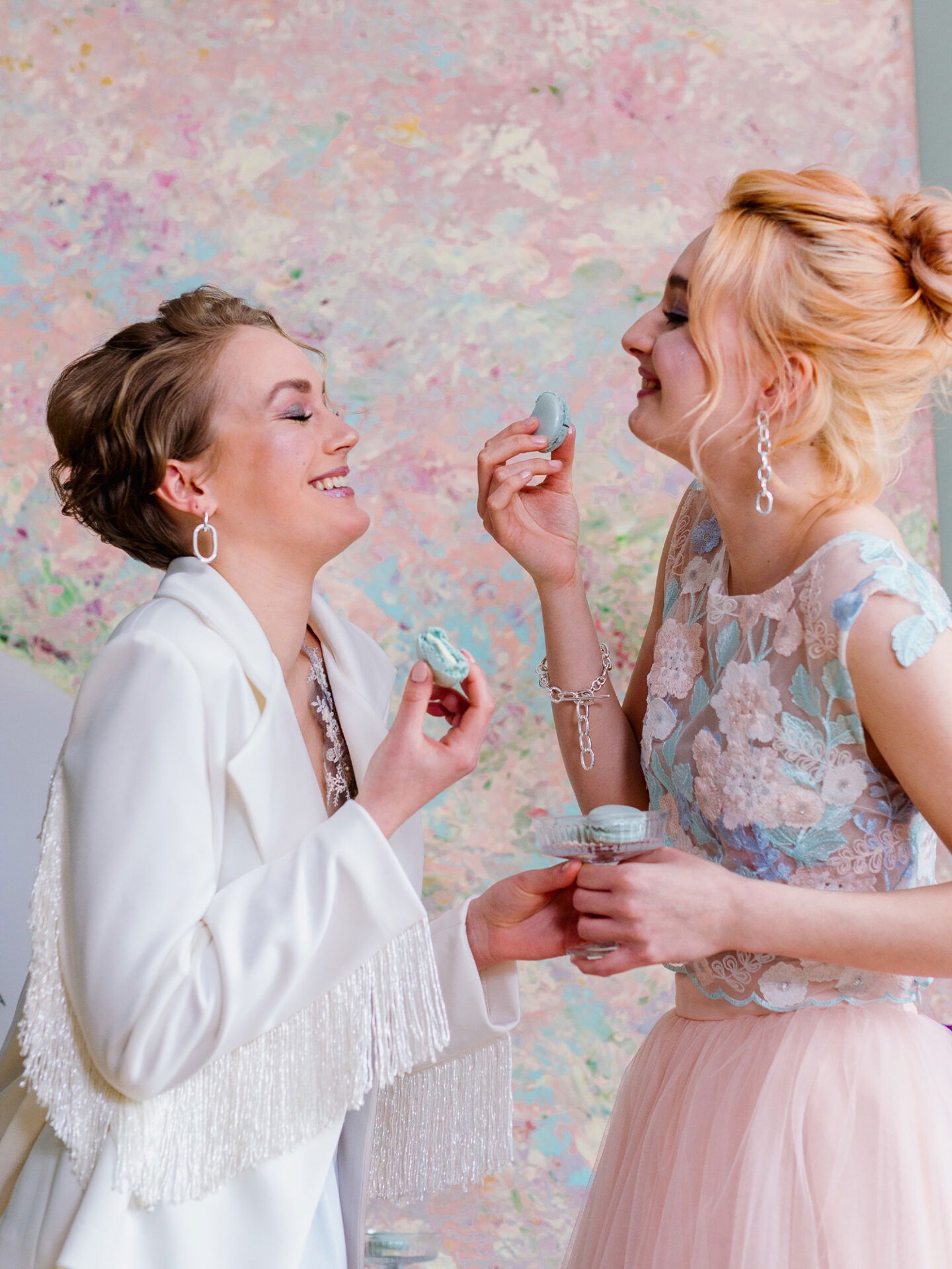 Die beiden Frauen lächeln sich an, während sie Macarons miteinander teilen. Eine trägt einen stilvollen Blazer mit langen Fransen, während die andere ein florales, pastellfarbenes Spitzenoberteil trägt. Im Hintergrund leuchtet eine künstlerische Wand in sanften Farben.