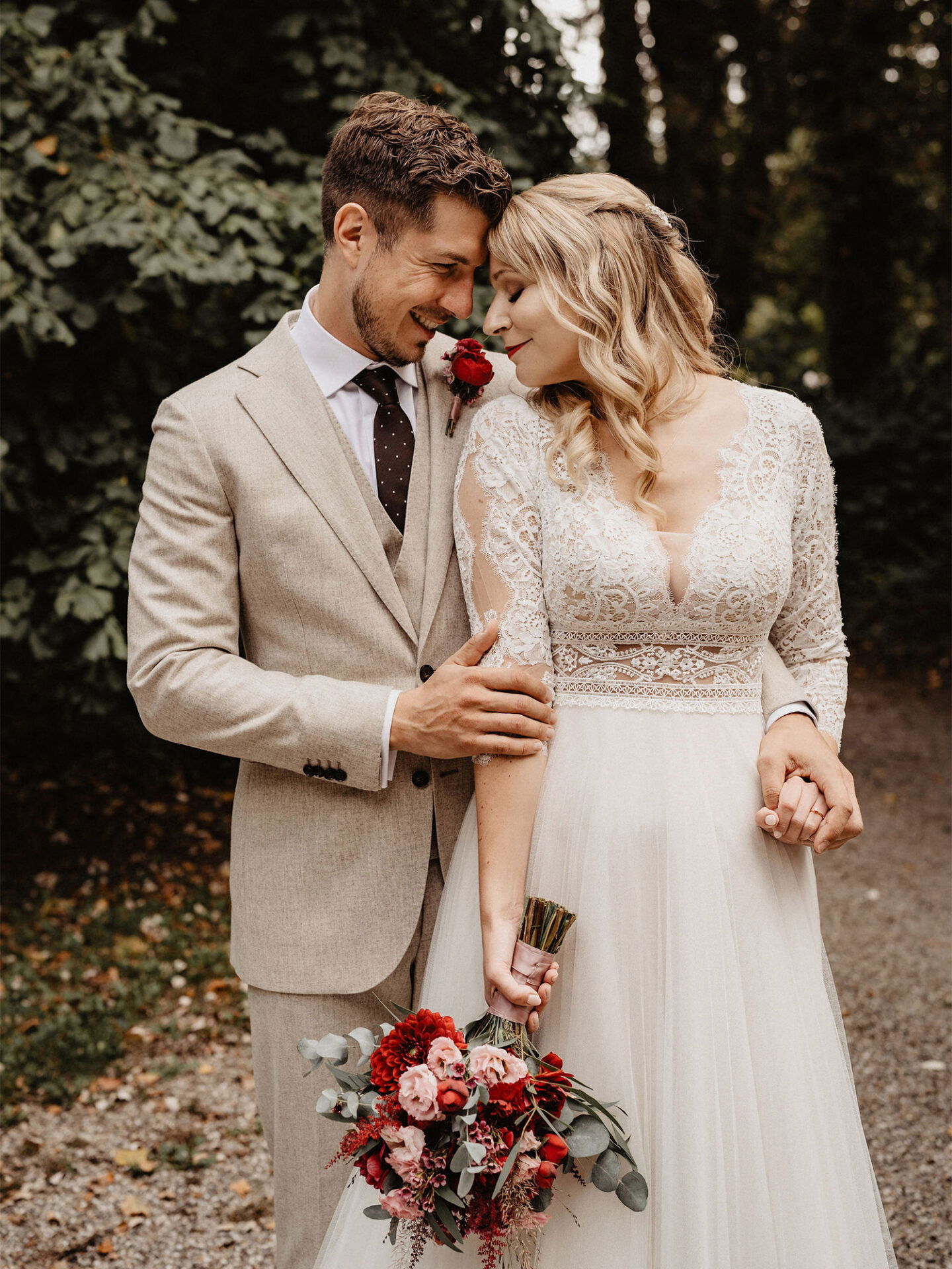 Ein Brautpaar steht in einer intimen Umarmung auf einem Waldweg. Die Braut trägt ein elegantes, langes Kleid aus Spitze und Tüll, und der Bräutigam trägt einen beigen Anzug. Sie hält einen bunten Blumenstrauß, und beide wirken glücklich.