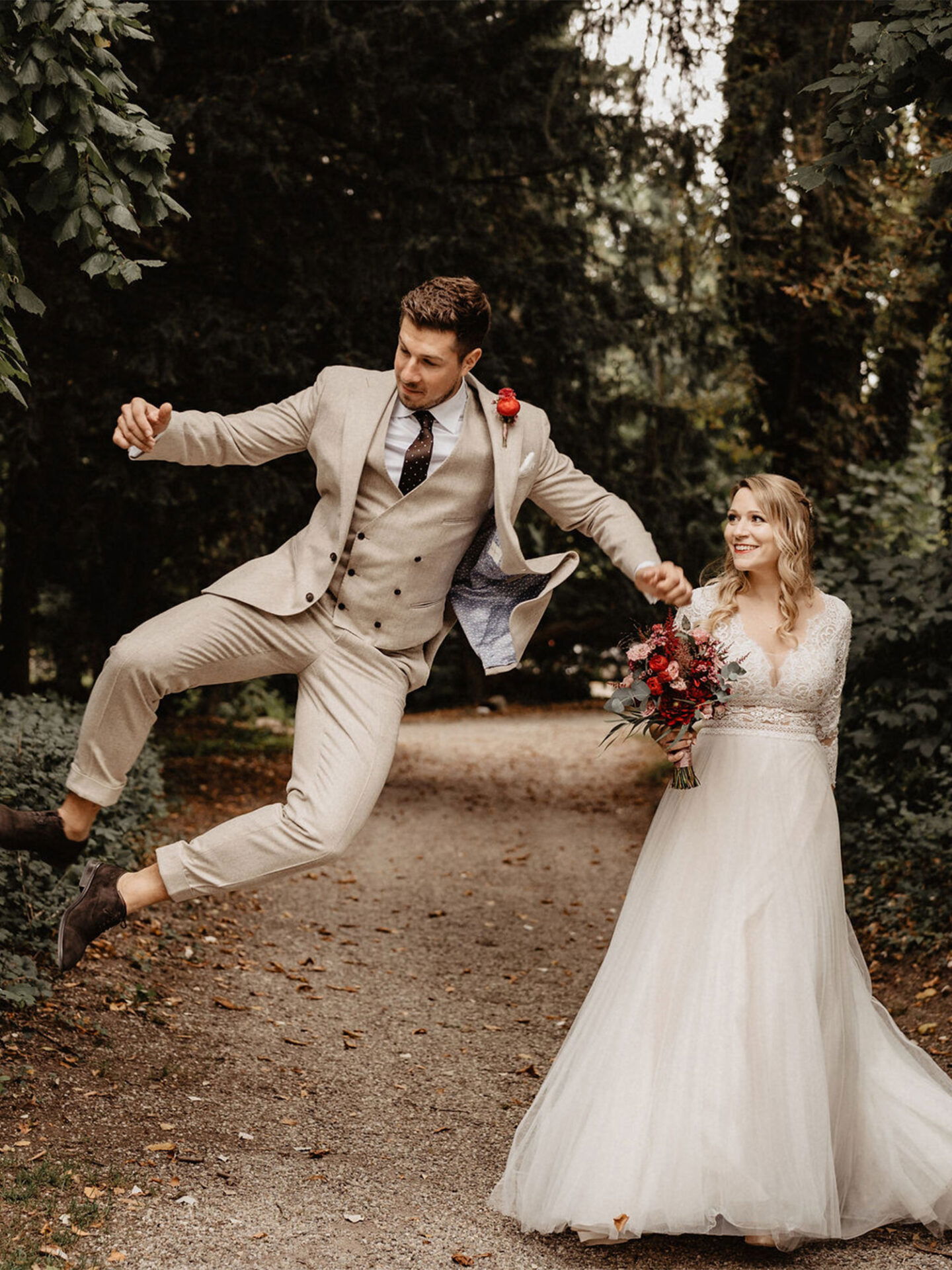 Ein witziger Moment während des Fotoshootings: Der Bräutigam springt hoch in die Luft, während die Braut ihn mit einem Strauß in der Hand an der Hand hält. Beide sind in Hochzeitskleidung und stehen auf einem schmalen Waldweg.