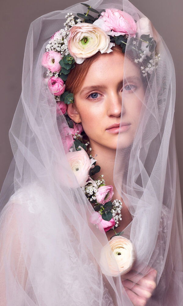 Eine Braut mit einem floralen Kranz aus Ranunkeln und Eukalyptus trägt einen luftigen Schleier, der ihr Gesicht verhüllt. Der Kranz aus rosa, weißen und cremefarbenen Blumen verleiht der Braut einen natürlichen, aber eleganten Look, während der Schleier eine geheimnisvolle Note hinzufügt.