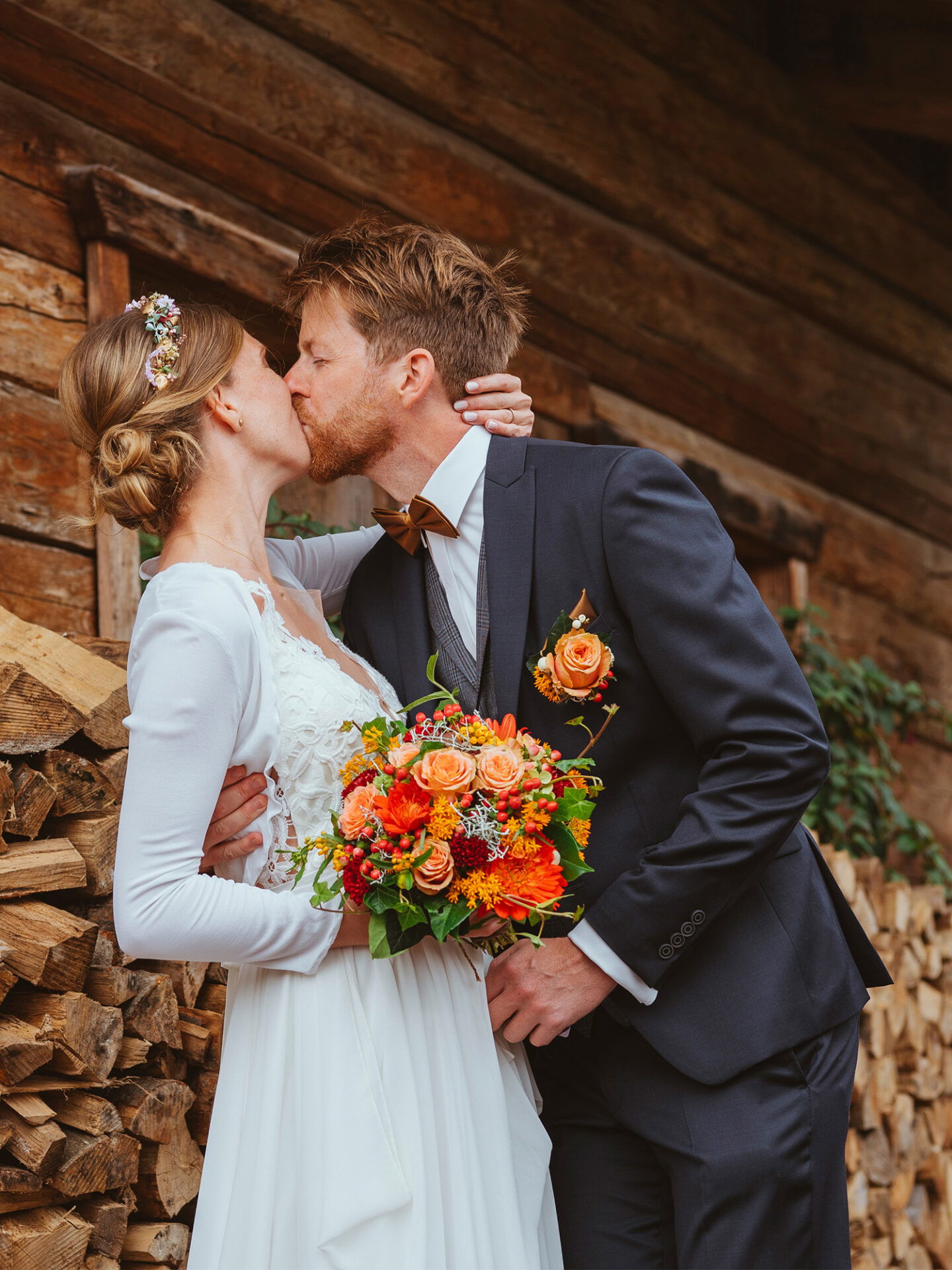 Das Brautpaar teilt einen Kuss vor einer rustikalen Hütte mit Holzscheiten im Hintergrund. Die Braut hat einen wunderschönen bunten Blumenstrauß, während der Bräutigam in einem klassischen Anzug mit Fliege steht.