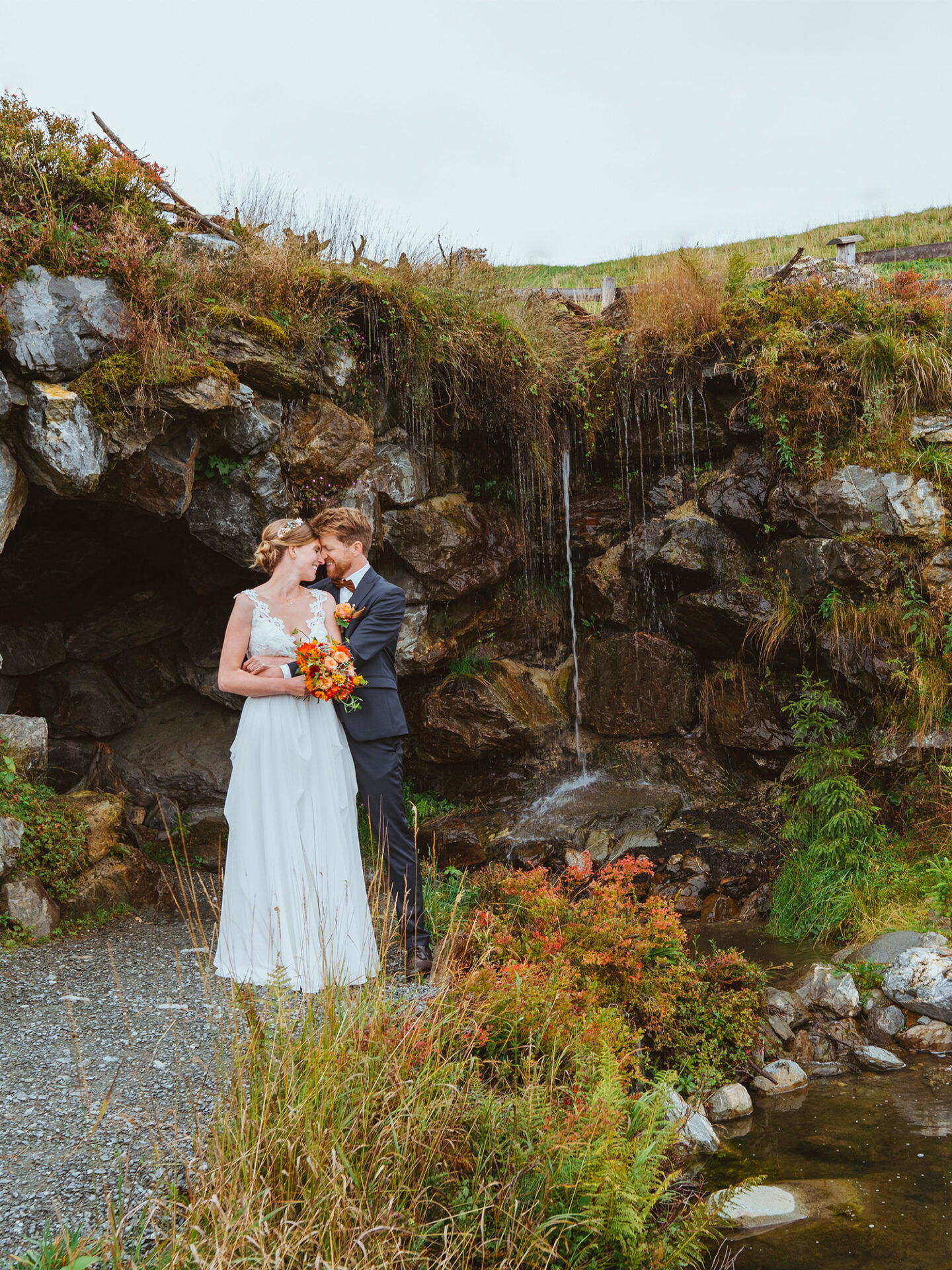 Das Paar steht vor einem Wasserfall, eingebettet in eine felsige Landschaft mit üppiger Vegetation. Die Braut hält einen Strauß orangefarbener Blumen, und die beiden lehnen die Stirn aneinander.