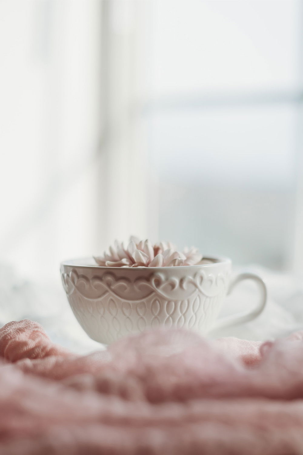 Eine weiße Teetasse mit filigranen Mustern ist mit einer Blume dekoriert und steht auf einem rosa Tuch. Das Bild strahlt Ruhe und Entspannung aus und lädt zu einem Moment der Achtsamkeit ein.