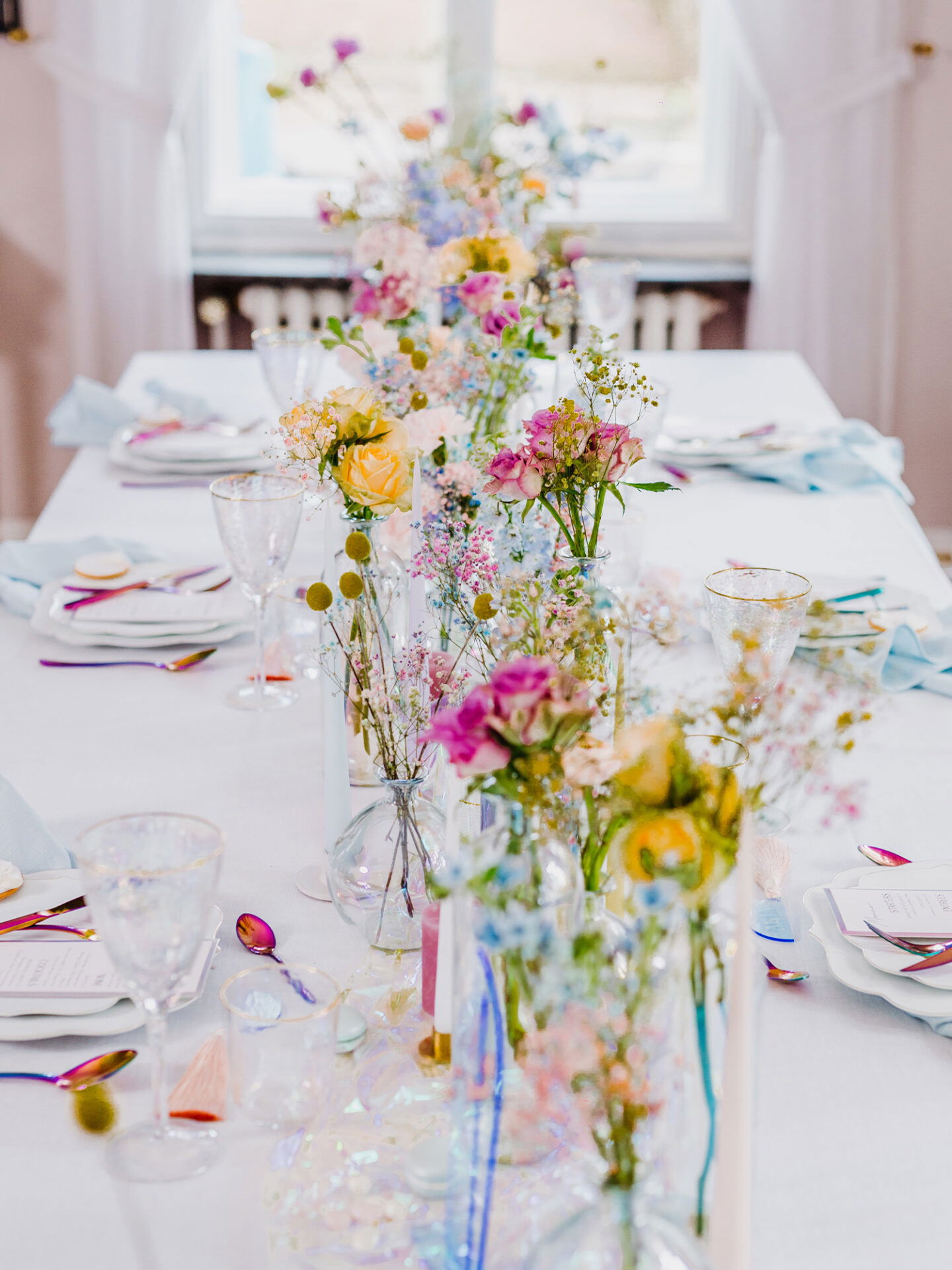 Der weiß gedeckte Tisch ist mit Glasvasen dekoriert, die zarte Blumen in Pastellfarben enthalten. Im Hintergrund lässt das Tageslicht den Raum erstrahlen. Das Arrangement wirkt elegant und festlich, mit farblich abgestimmtem Besteck und Glaswaren.