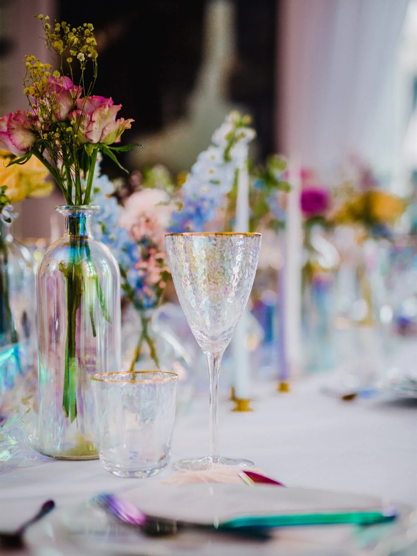 Elegante Tischdekoration mit einem goldverzierten Weinglas und einem kleineren Glas, die beide in einem irisierenden Licht schimmern. Daneben steht eine Vase mit rosafarbenen Blüten, die der festlichen Szenerie einen romantischen und frischen Touch verleihen.